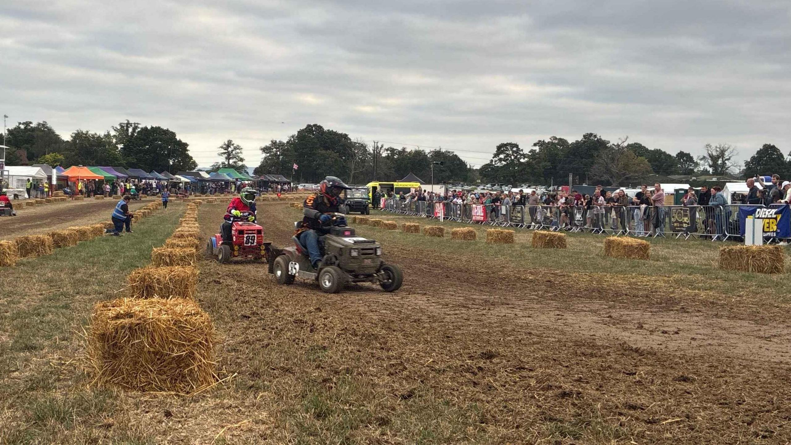 Two men on lawn mowers with racing colours and numbers racing down a a dirt track