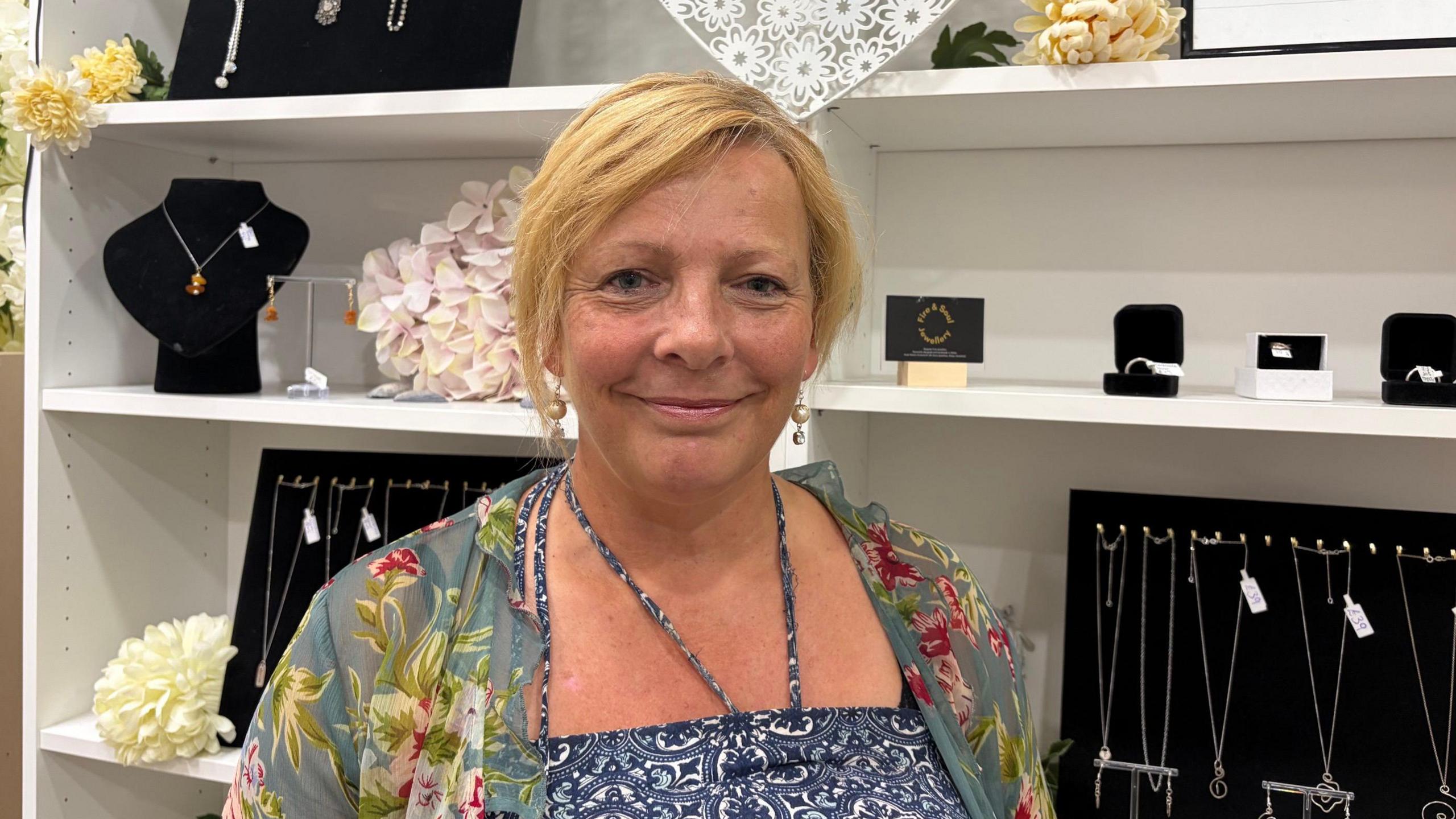 Heidi in a blue and white patterned dress and a patterned green and pink cardigan standing in front of jewellery. She has short blonde hair and smiles at the camera.