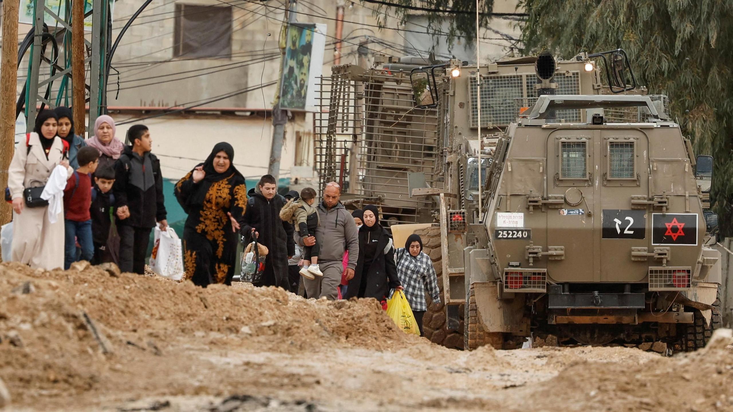 Palestinians walk next to Israeli military vehicles and an armoured bulldozer on a destroyed road from Jenin refugee camp, during an operation by Israeli security forces, in the occupied West Bank (22 January 2025)