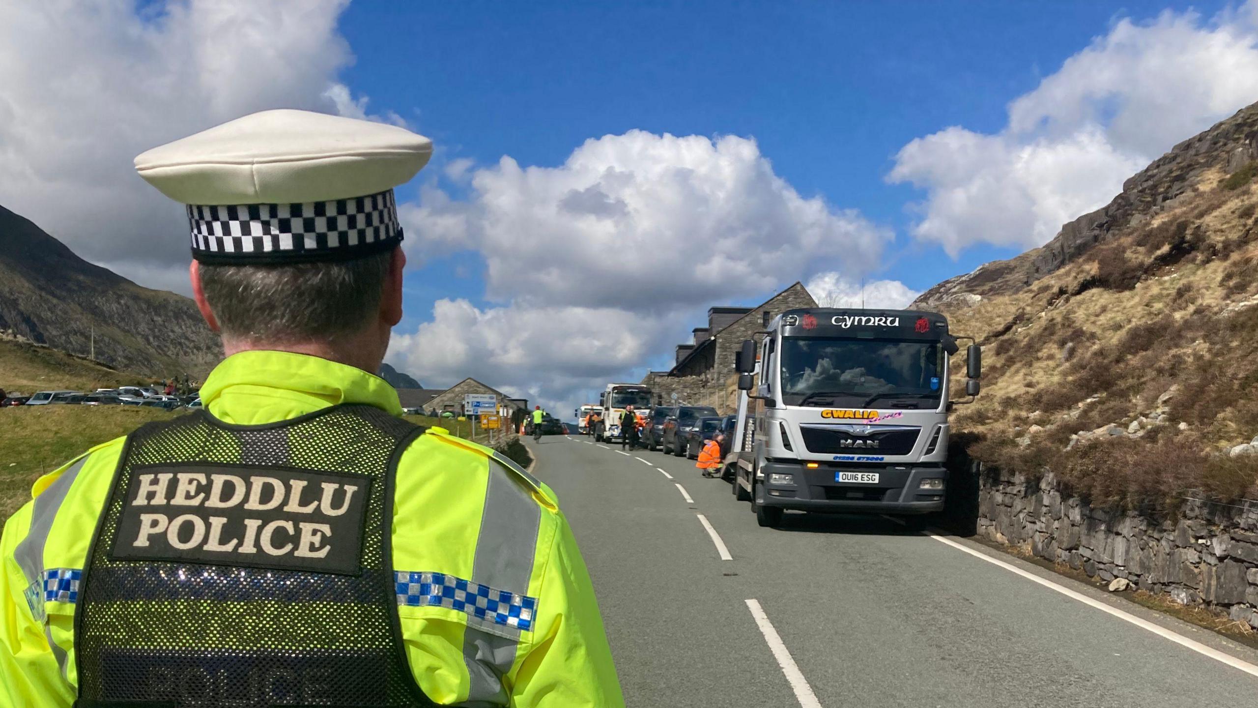 Tow trucks at Pen y Pass A4086, Nant Peris, Caernarfon, Gwynedd in April 2023 with a police officer in the forefront, dressed in uniform and facing away from the camera 