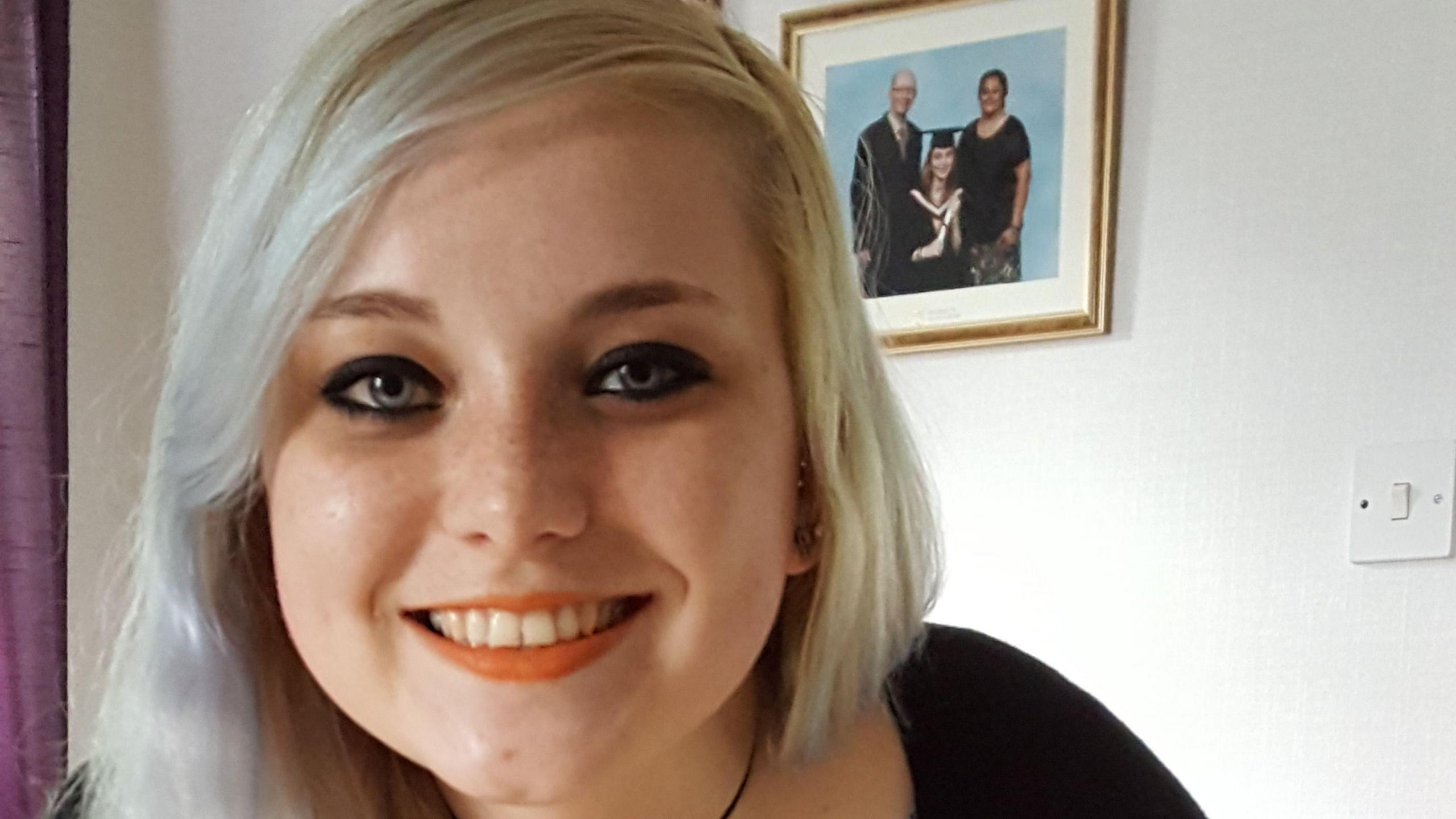 A smiling woman with blonde hair looking directly at the camera with a picture of her own graduation behind her