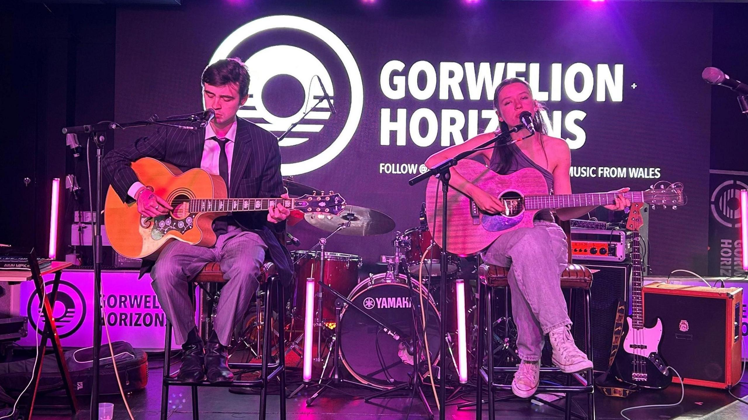 Mae Nancy a Harry yn perfformio ar y llwyfan. Mae gan y ddau gitarau ac maen nhw'n canu i'r standiau meicroffon. Y tu ôl iddyn nhw ar sgrin fawr mae poster Gorwelion Horizons. Mae Nancy, ar y dde, yn gwisgo jîns glas, top brown oddi ar yr ysgwydd ac esgidiau hyfforddi gwyn. Mae Harry, ar y chwith, yn gwisgo trowsus llwyd, crys gwyn, tei a siaced. Mae amryw o offerynnau a gwifrau o'u cwmpas ar y llwyfan.