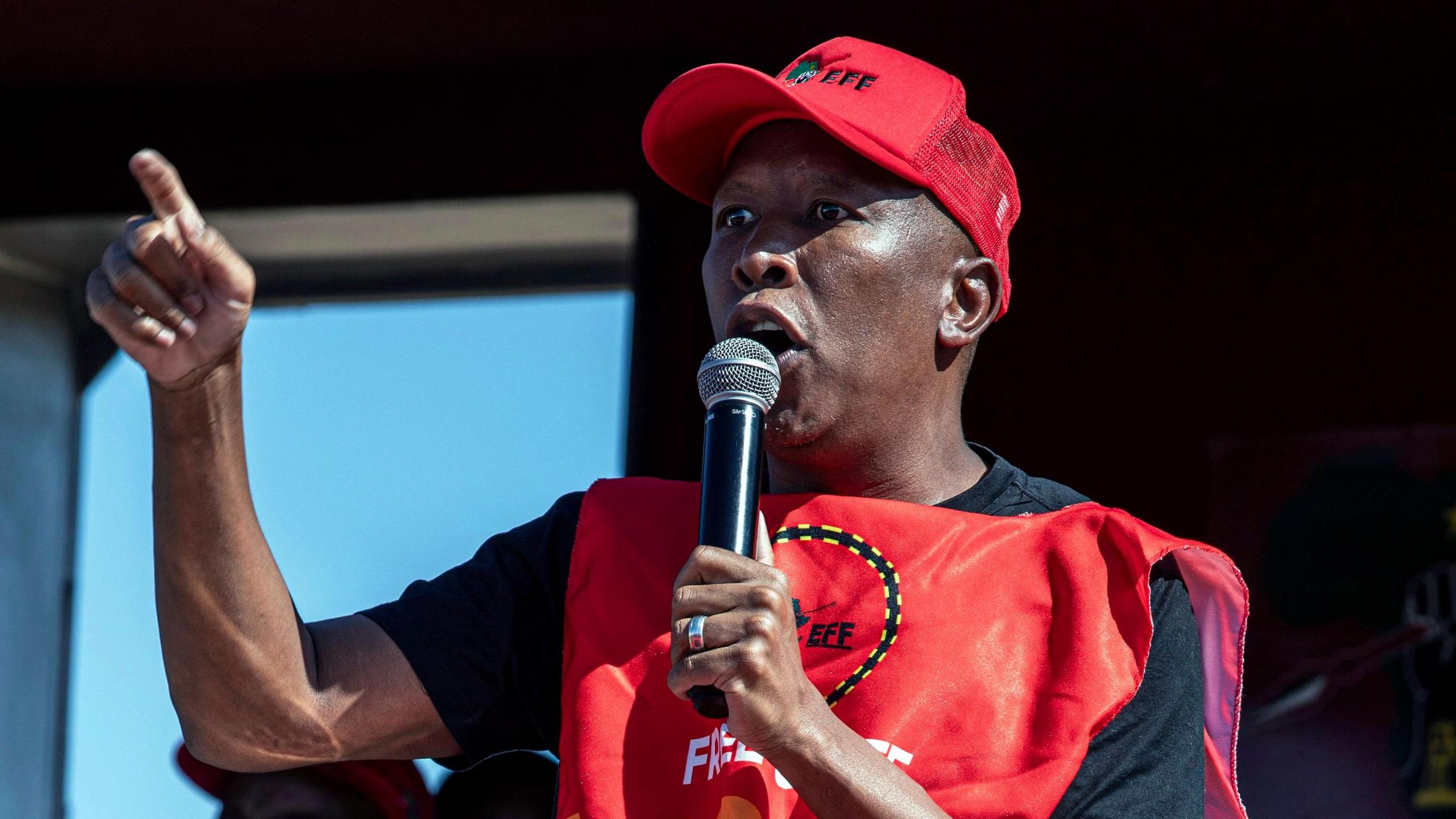 Julius Malema, wearing a red Economic Freedom Fighters-branded cap and vest, speaks into a microphone and gesticulates with his right arm on stage at a political rally