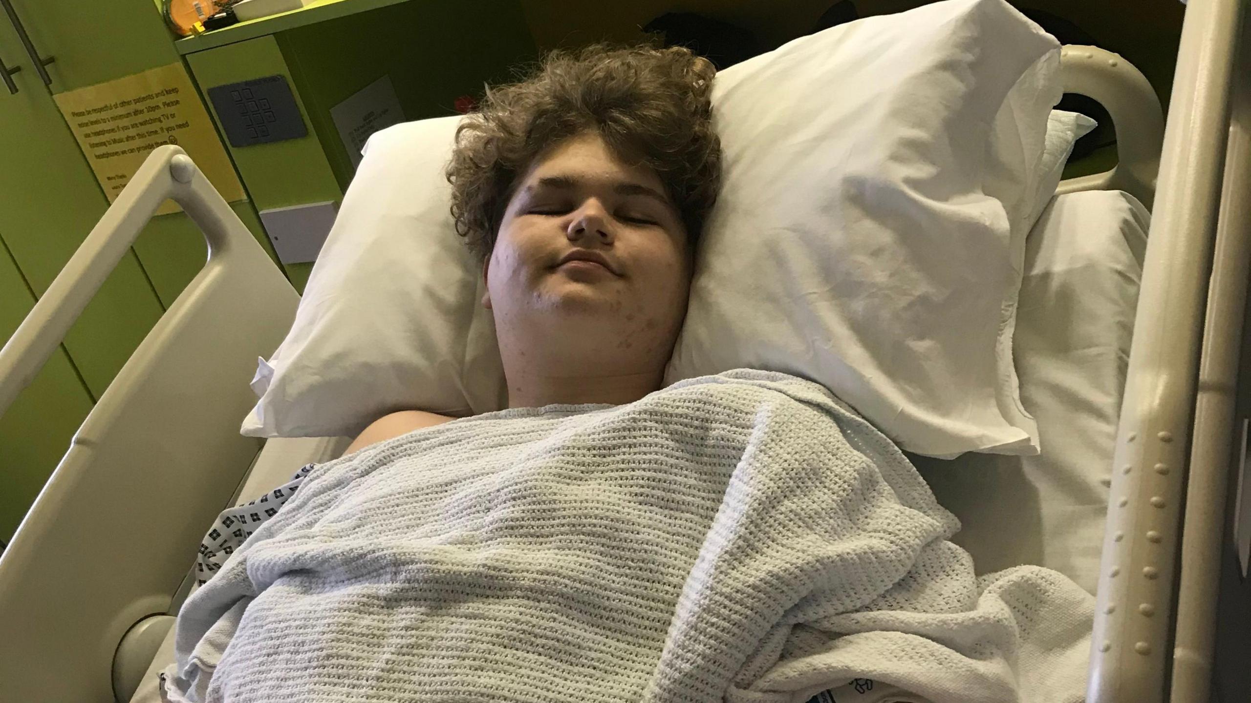 A teenager lying on a hospital bed earing a hospital gown and under a blanket. He appears to be sleeping.