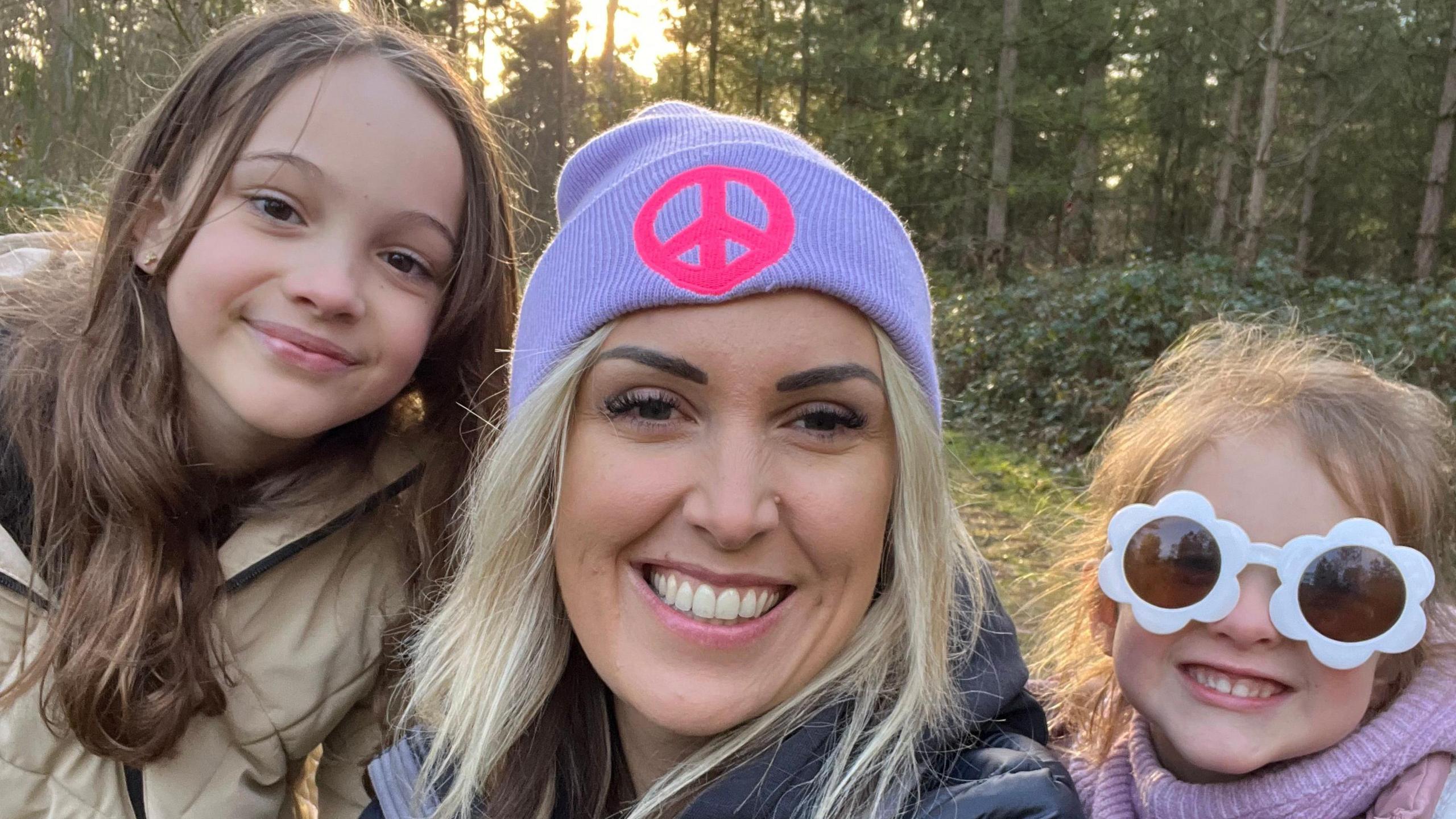 Keri Bird doing a smiling selfie with her two young daughters, outside in a forest. Keri has long blonde hair and is wearing a lilac woolly hat with a pink 'peace' symbol on the front, and a navy coat. The daughter on the left has long brunette hair and a beige coat. The girl on the right is wearing big white flowery sunglasses and a lilac woolly jumper underneath a pink coat.