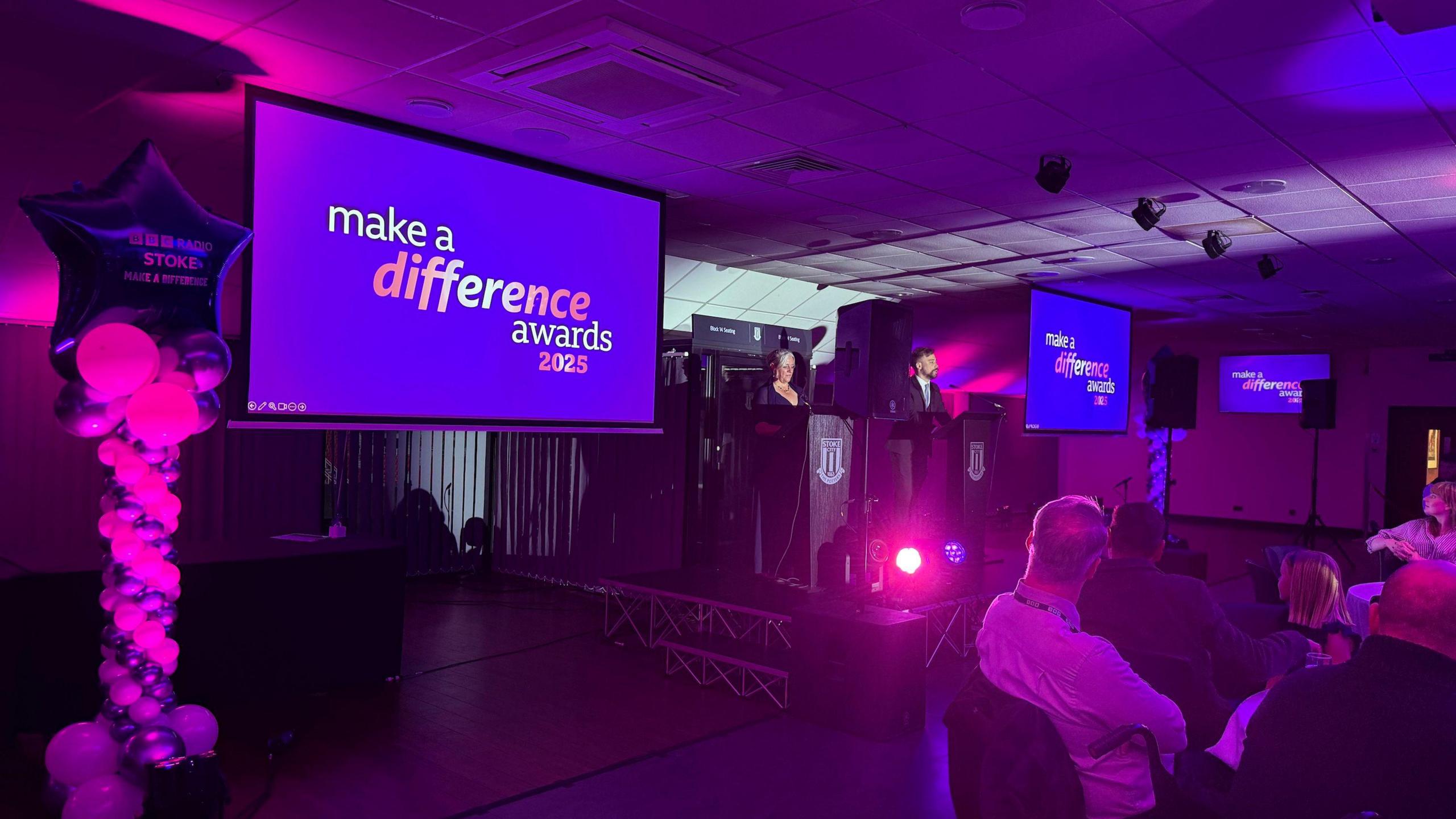 Jodie Looker and Lee Blakeman, in the centre of the image, are stood at podiums. Either side of them are large tv screens with purple BBC Radio Stoke Make A Difference Awards branding. There is pink lighting and members of the audience are watching the stage.