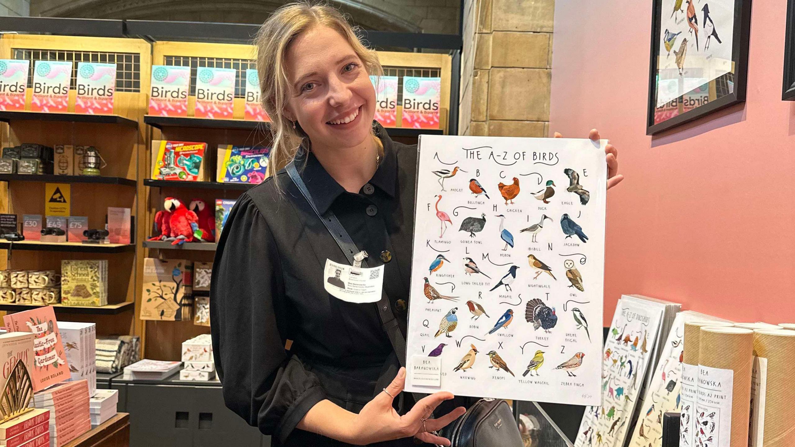 Bea Baranowska holding a print of The A-Z of Birds. It features her drawings of various species. Bea has blonde hair which is tied up and is wearing a black collared shirt with an ID pass attached to it. She is smiling at the camera. 