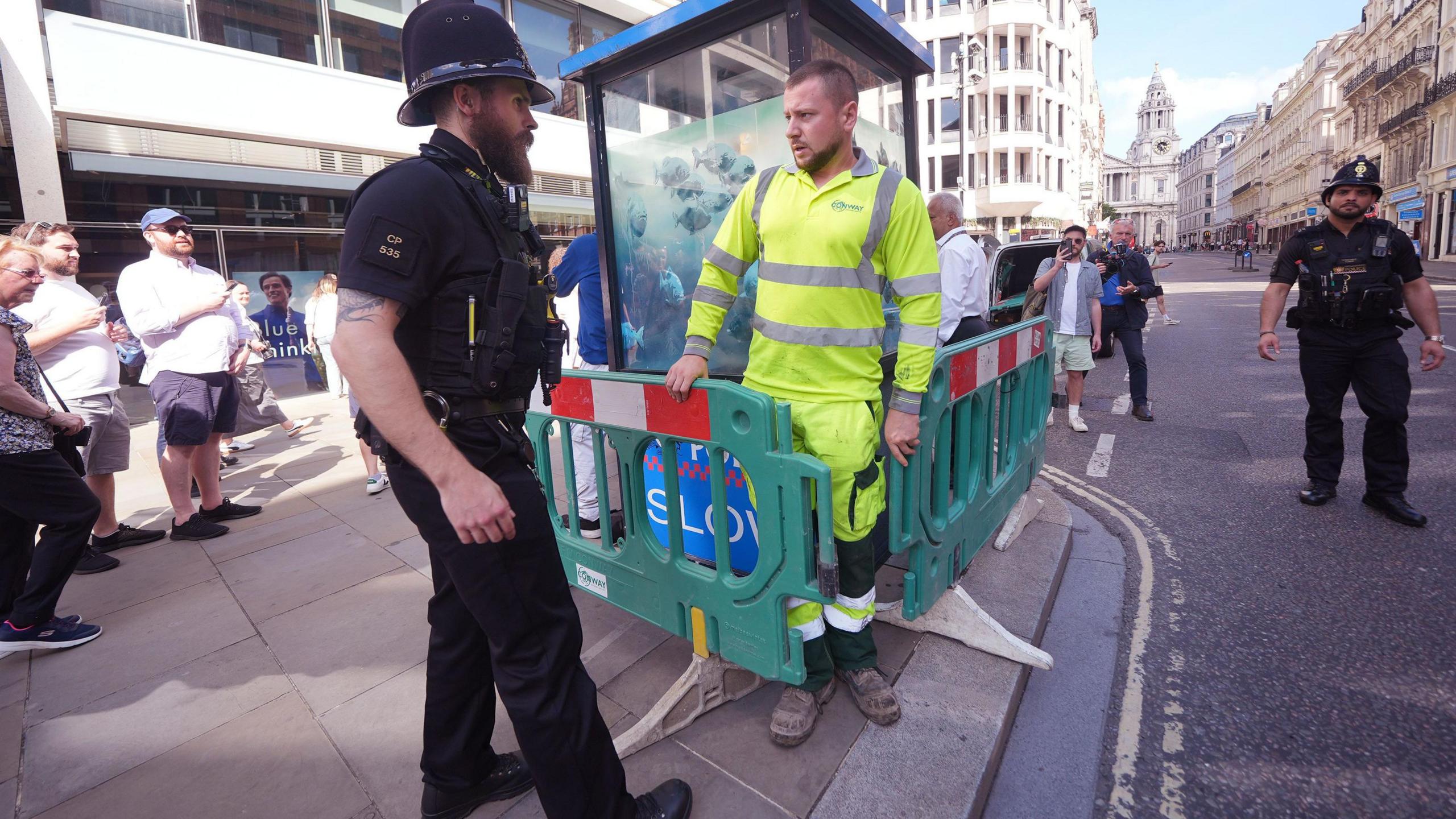 Contractor wearing high vis top and trousers erecting barrier round box while a police officer stands by. Passers by are watching the scene from the pavement.