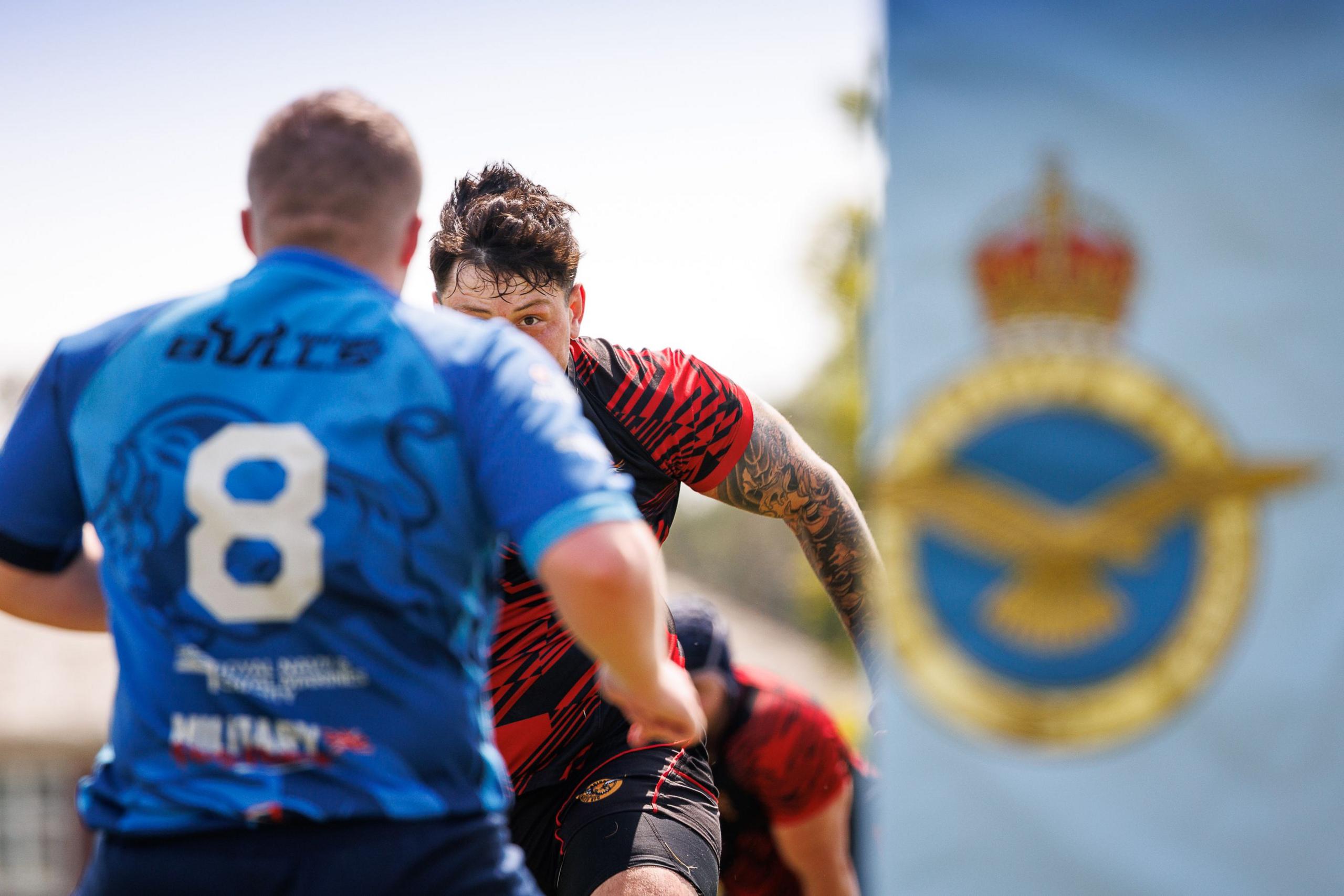 Two rugby players on opposing sides rush at each other.  The player facing the camera has his face mostly covered by his opponent's shoulder, but his eye is visable.