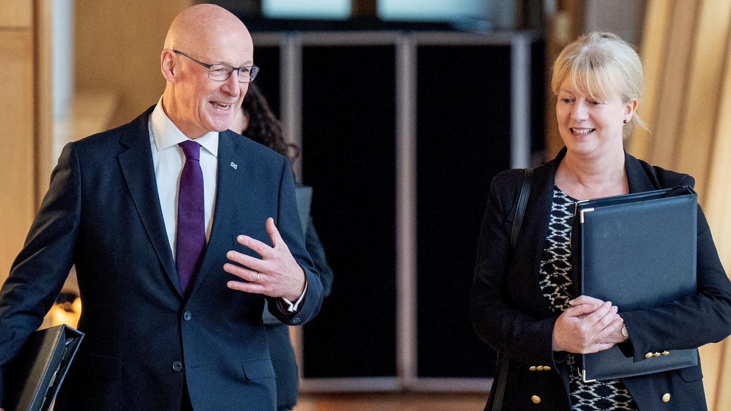 John Swinney and Shona Robison walking along a corridor in the Scottish Parliament. He is smiling and has his left hand in front of him, while she is holding a black folder. 