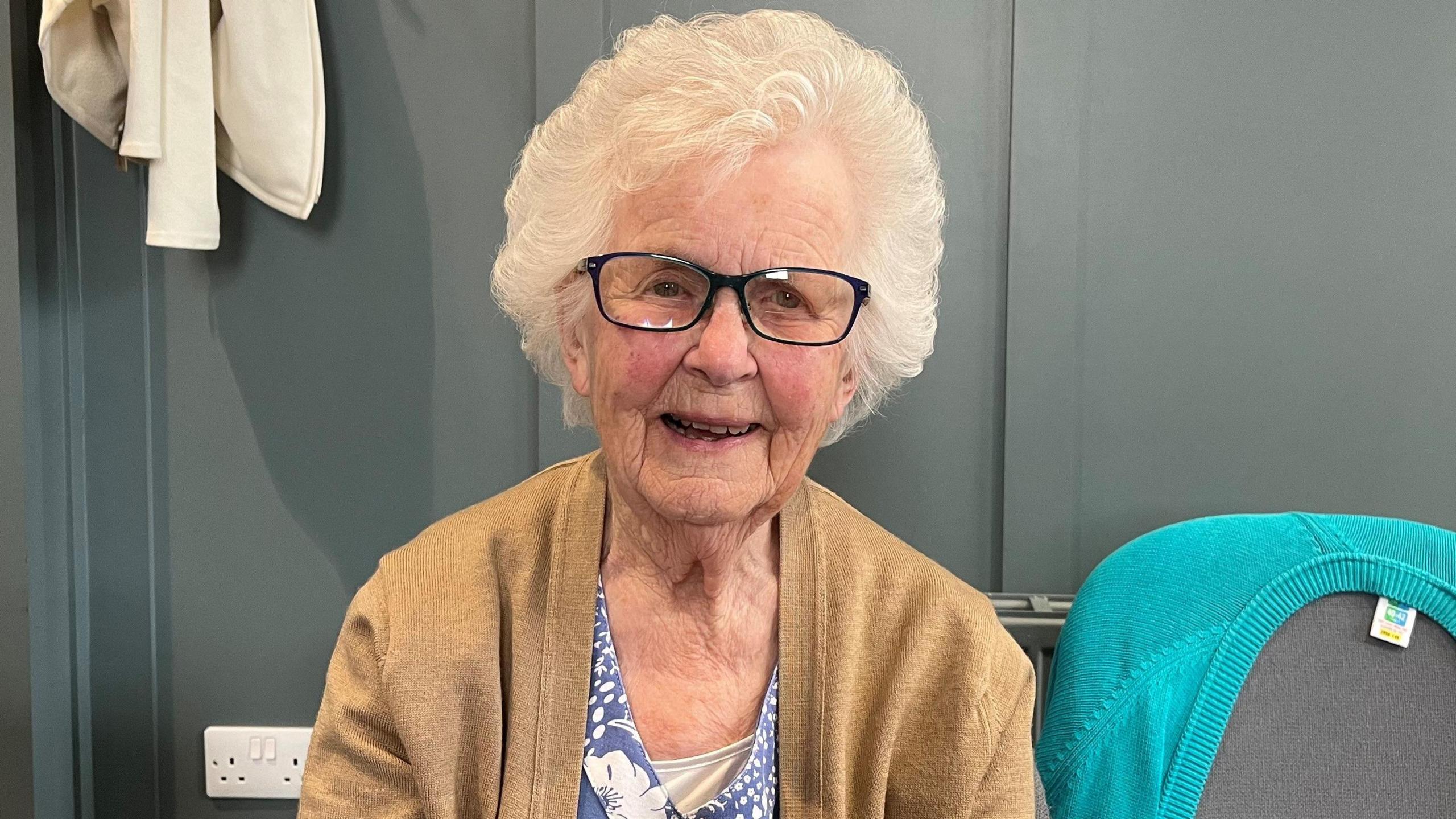 A photo showing the head and shoulders of an elderly white woman, She is wearing glasses and has white hair. She is sat down in front of a dark wall