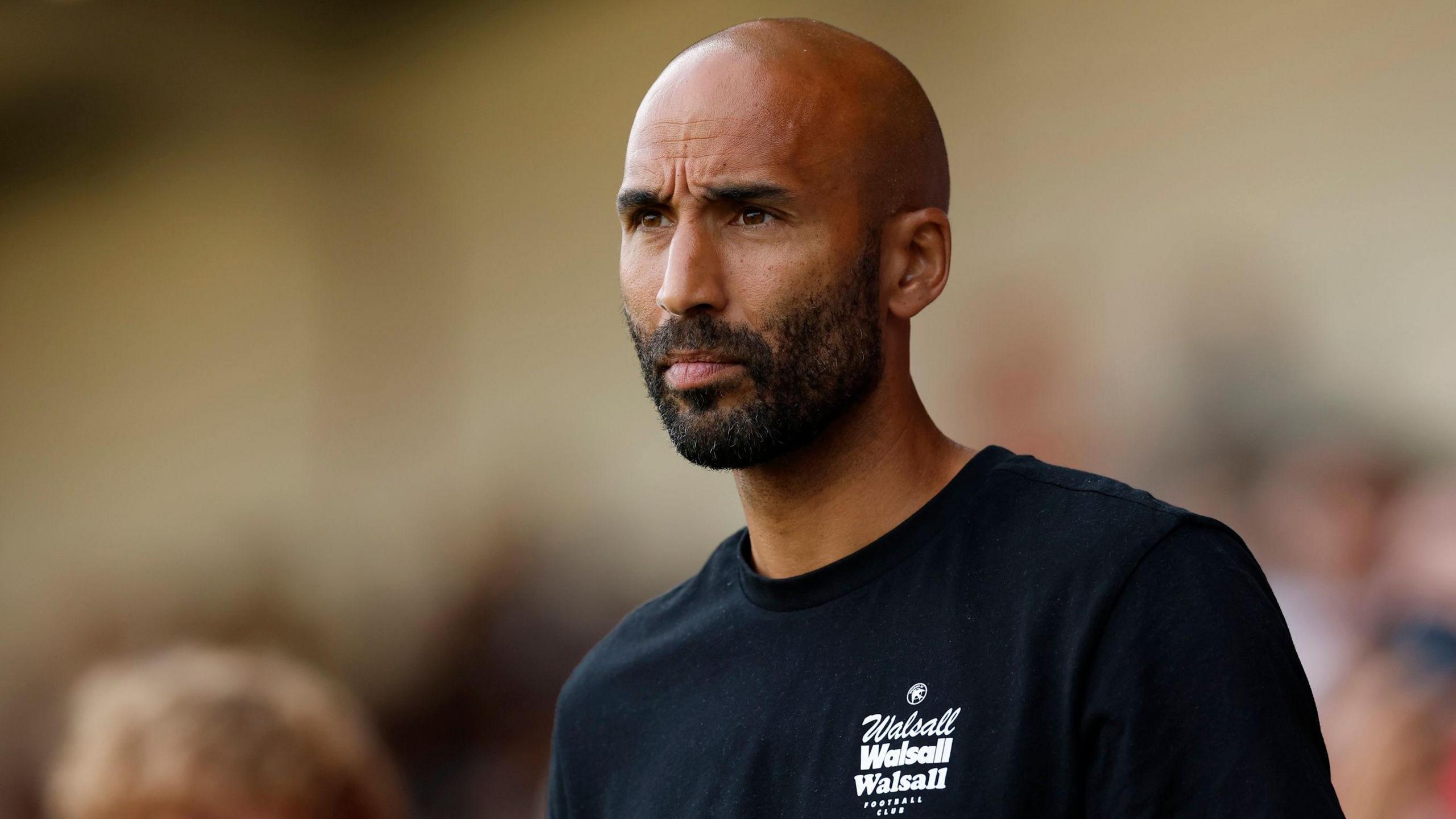 Walsall head coach Lee Grant watches his side from the touchline. He is wearing a dark sweatshirt with Walsall written in white on front.