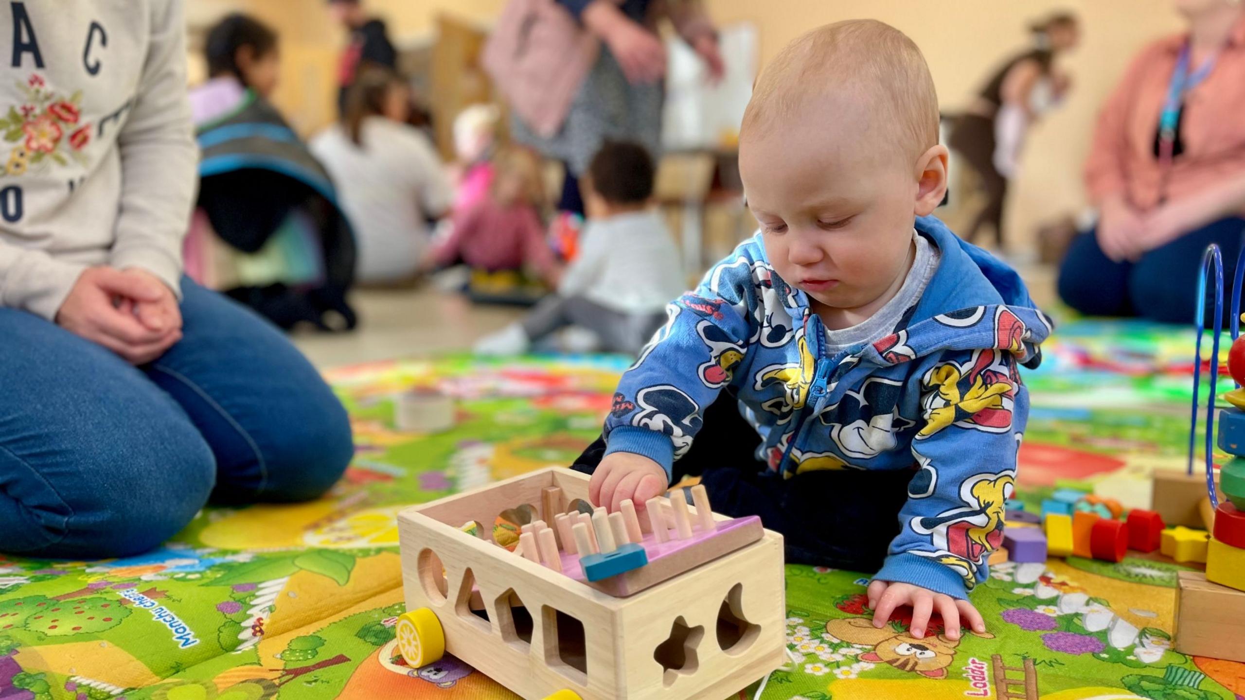 A toddler in a blue zipped hoodie is on a playmat playing with a wooden toy. The toddler is wearing black trousers, a grey t-shirt and his hoodie is decorated with images of Mickey Mouse and Pluto the dog. The toy has yellow wheels and shapes cut out of the side. He has colourful wooden shape blocks next to him too. A woman sits beside him wearing blue jeans and a beige jumper with flowers on it. More children and adults can be seen blurred out in the background.