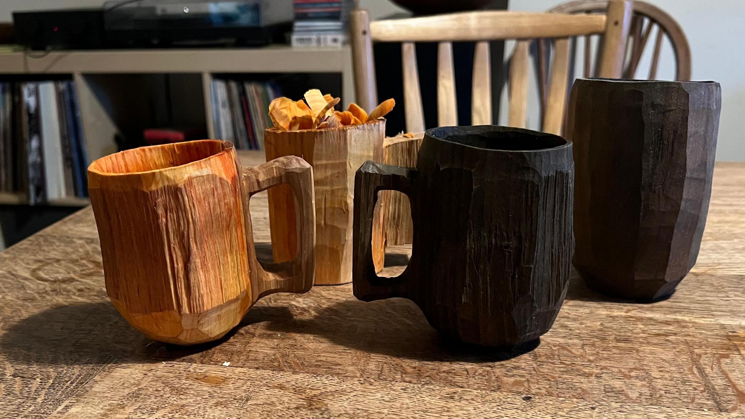 Four hand-carved wooden mugs in different shades - light brown, dark brown, and orange-brown - are placed on a wooden table. The background features a wooden chair and shelves filled with books or records, suggesting a cosy indoor setting.