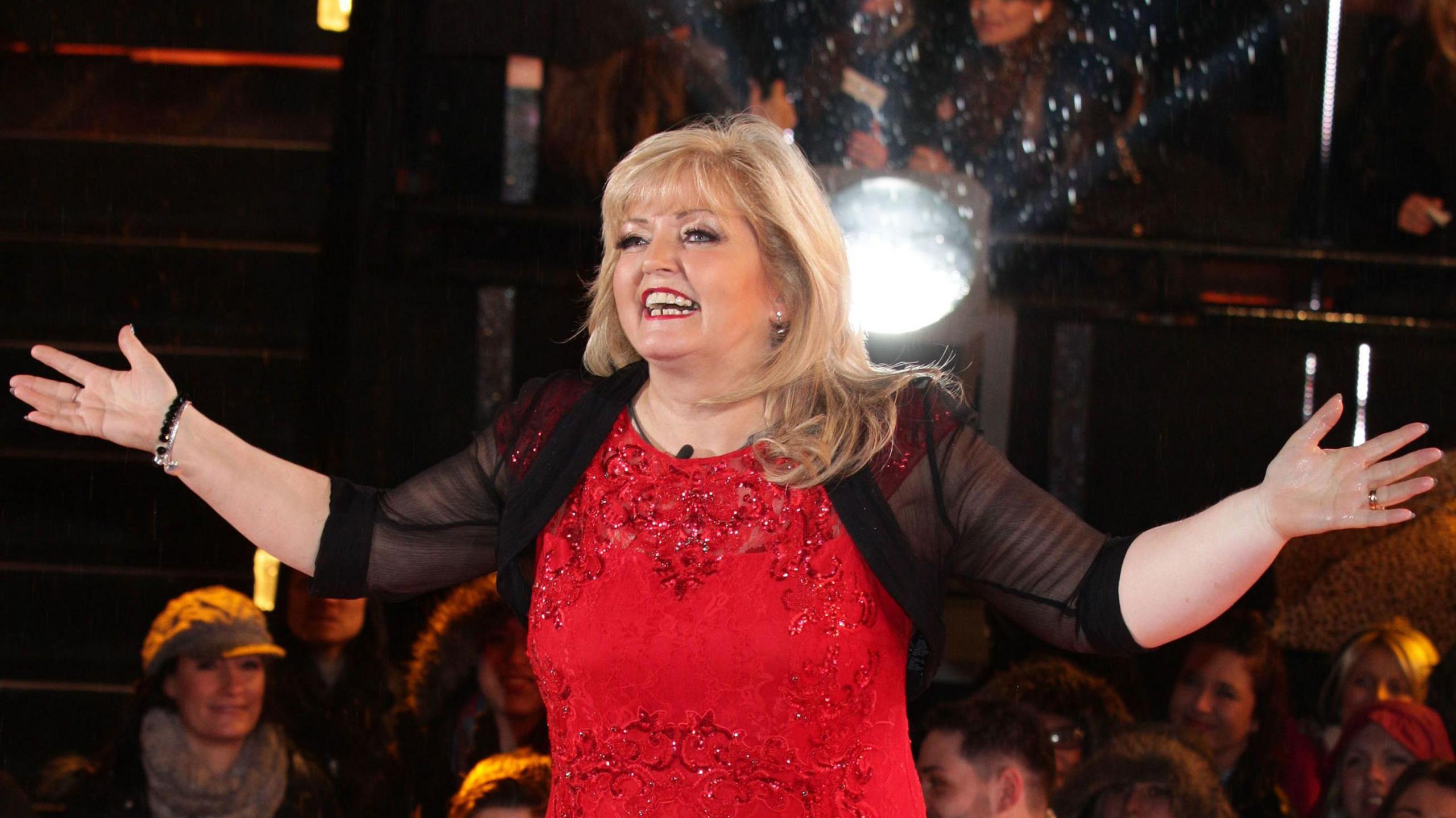 Linda Nolan, wearing a red dress, smiles at the camera with her arms outstretched.