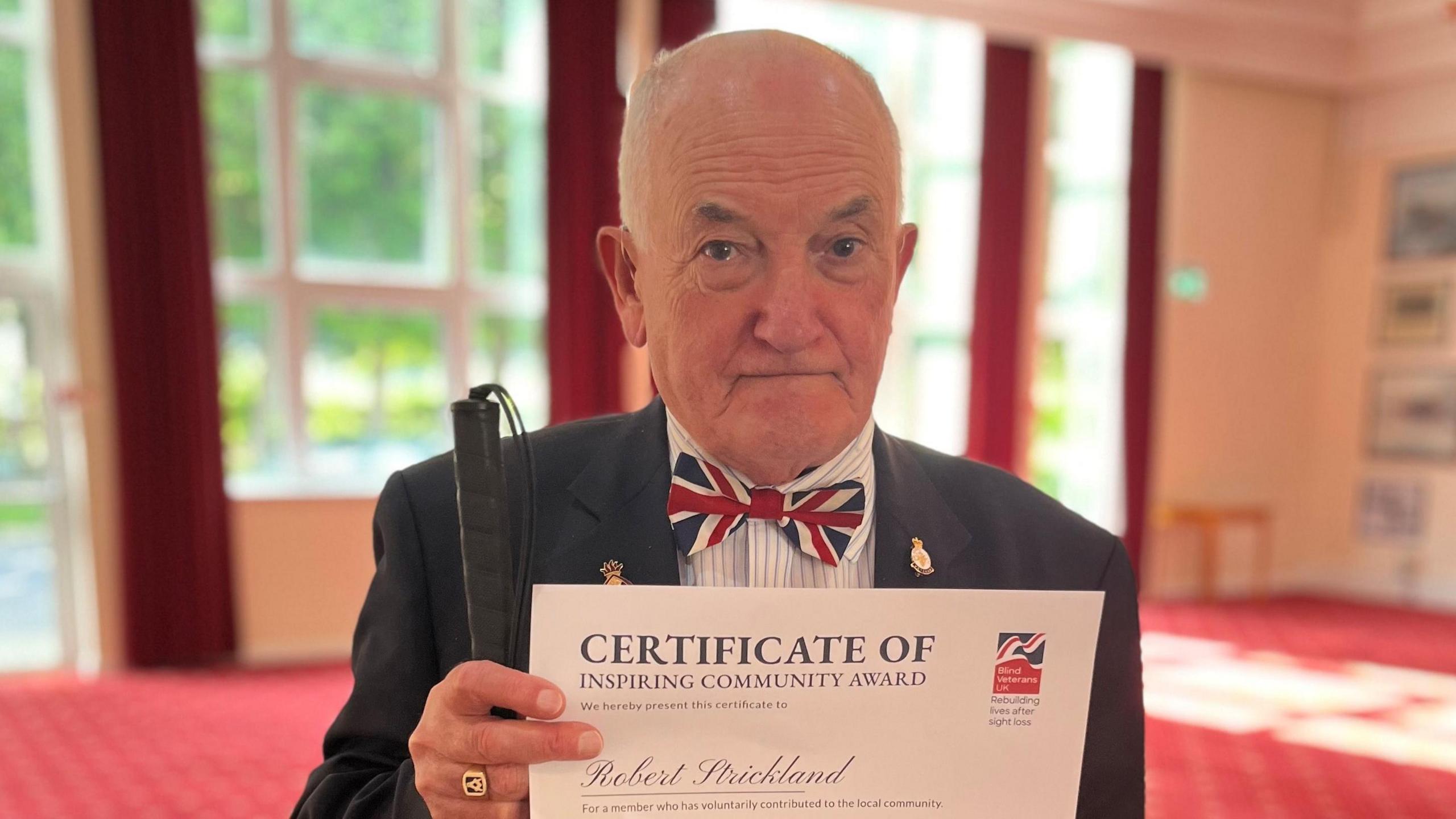 Bob Strickland wearing a union flag bow tie and a navy suit. He has a couple of gold badges pinned to the jacket. In his hand, he is holding a white cane with a black handle. He is also holding a certificate.