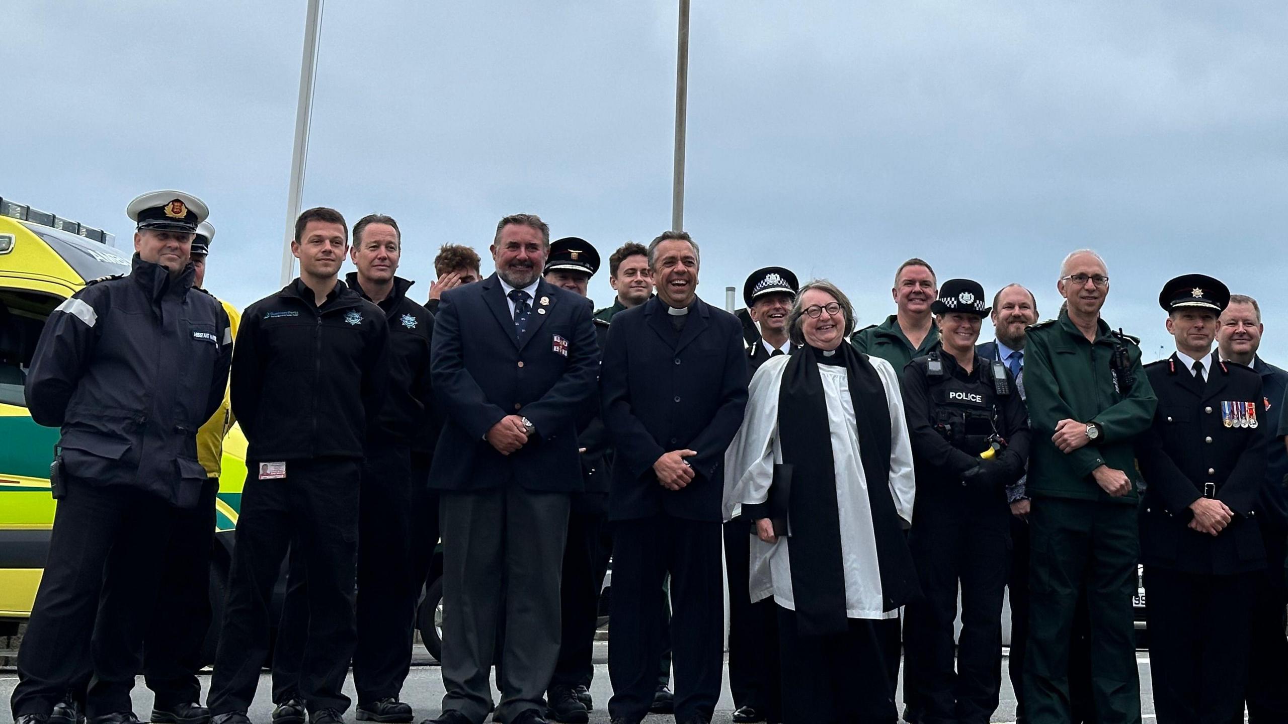 Men and women in different uniforms, including police, fire, paramedics and ecclesiastical, stand with hands clasped in front of two flag poles.