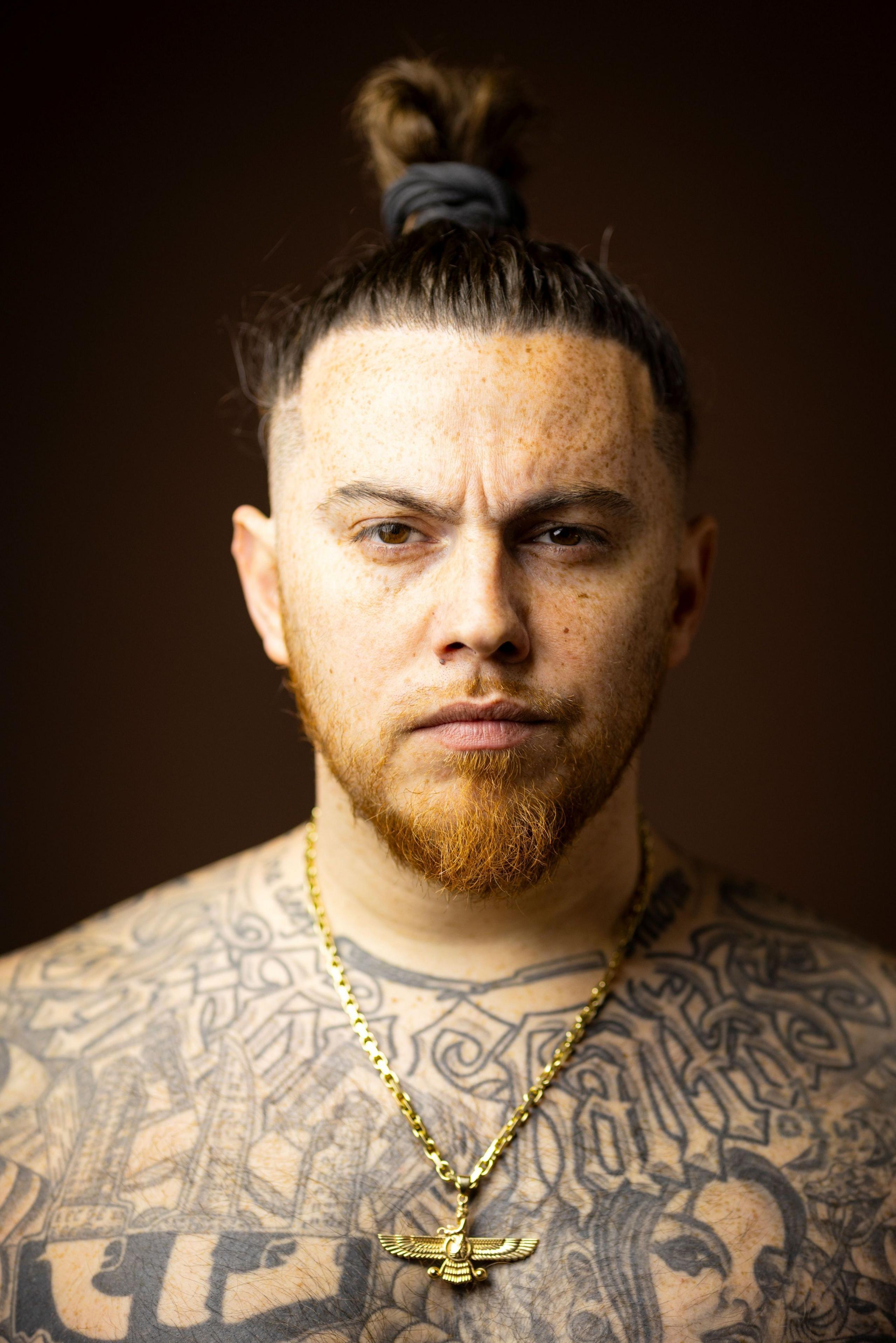 Close-up studio portrait of a person with hair tied in a top knot, freckles, a short beard, and extensive black-and-grey tattoos across the chest and shoulders. The person wears a gold chain necklace with a winged pendant and faces the camera against a dark brown blurred background.