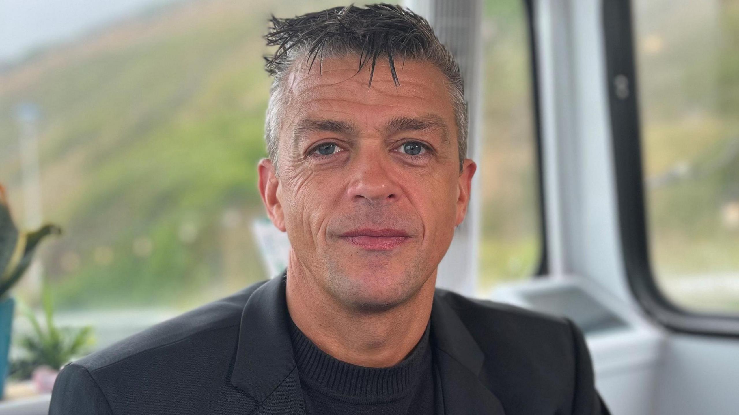 Lorne Castle smiles at the camera. He has gelled, grey hair which is short at the sides. He wears a dark jacket over a black, collarless top.