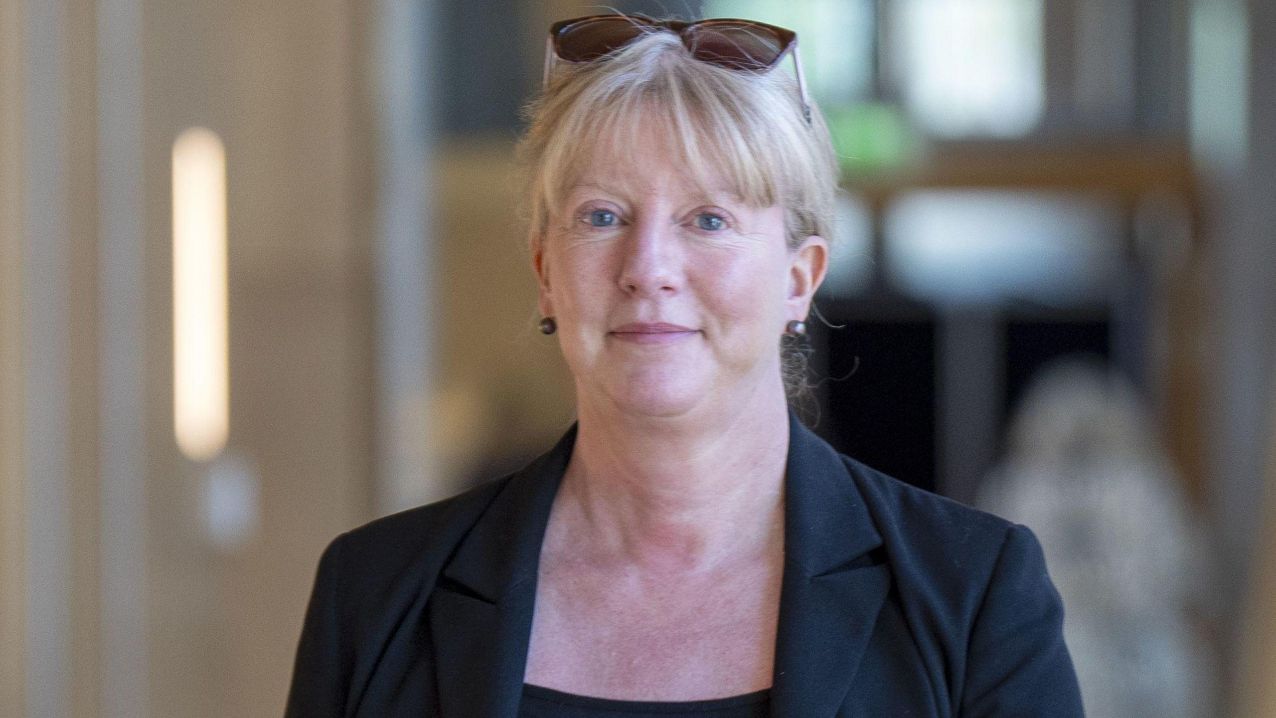 Head and shoulders shot of Shona Robison, a lady with tied back blonde hair, walking along a brightly lit corridor in the Scottish Parliament building in Edinburgh. She is looking directly at the camera and is slightly smiling. She is wearing a black top, matching jacket and has a pair of sunglasses balancing on her head.