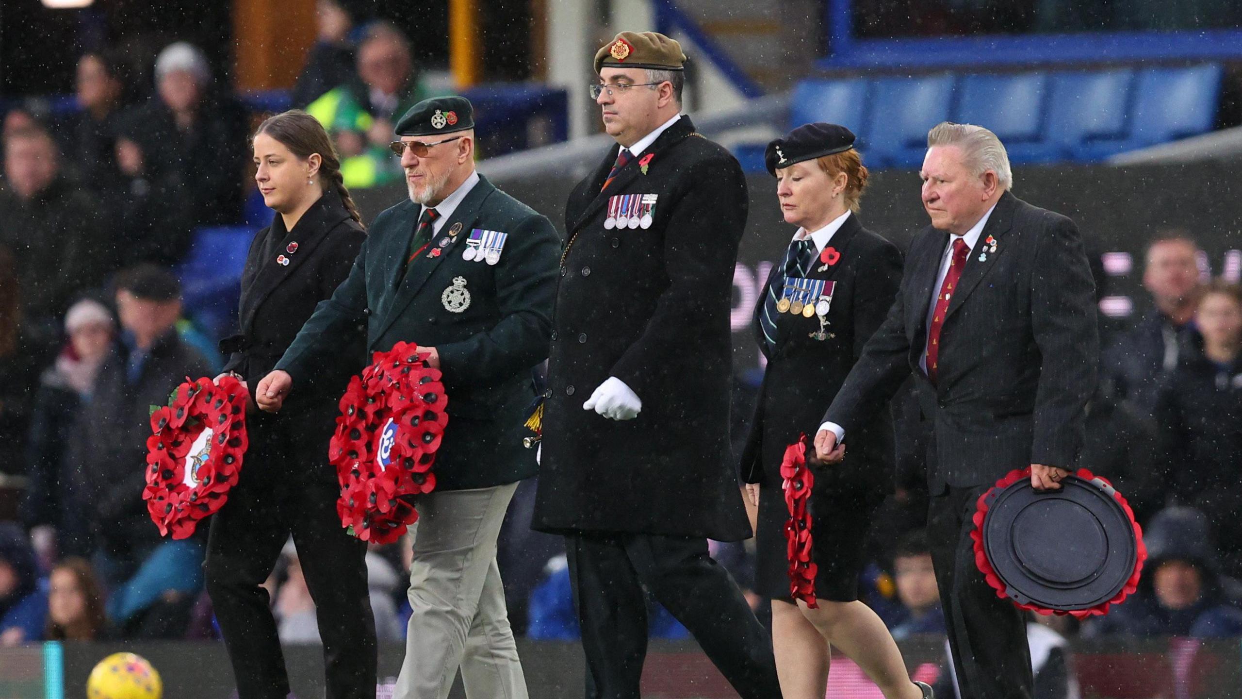 Five military veterans march out in the rain before the game in uniform and wearing medals on their lapels. They carry red poppy wreaths to lay on the pitch.