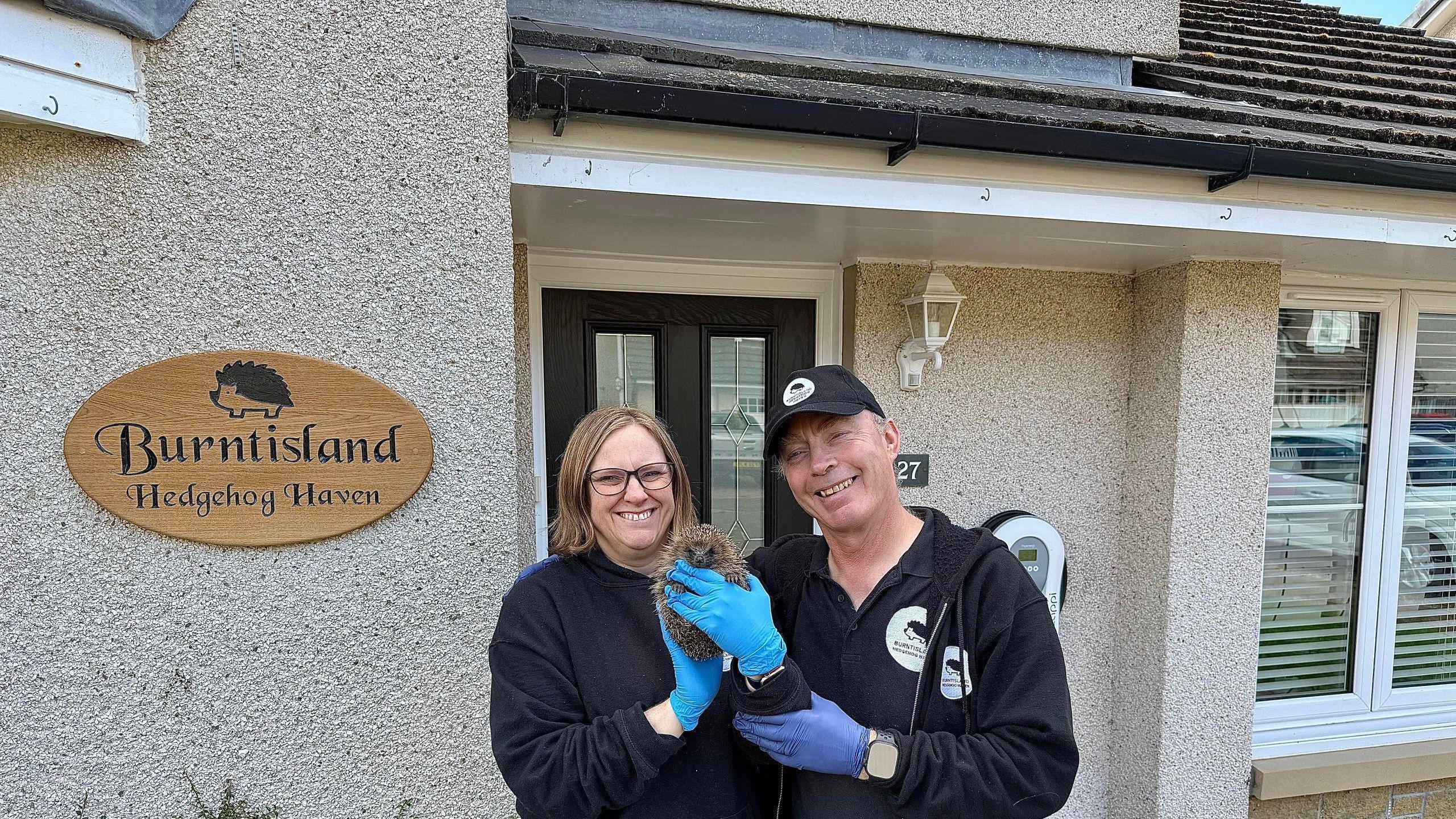 Sharon holds a hedgehog. She is wearing blue rubber gloves. Her husband Andy is standing beside her and smiling. He is wearing a black baseball cap and a dark top. She is wearing a dark jumper and is also smiling. They are standing in front of their house which has a sign on it that says Burntisland Hedgehog Haven.