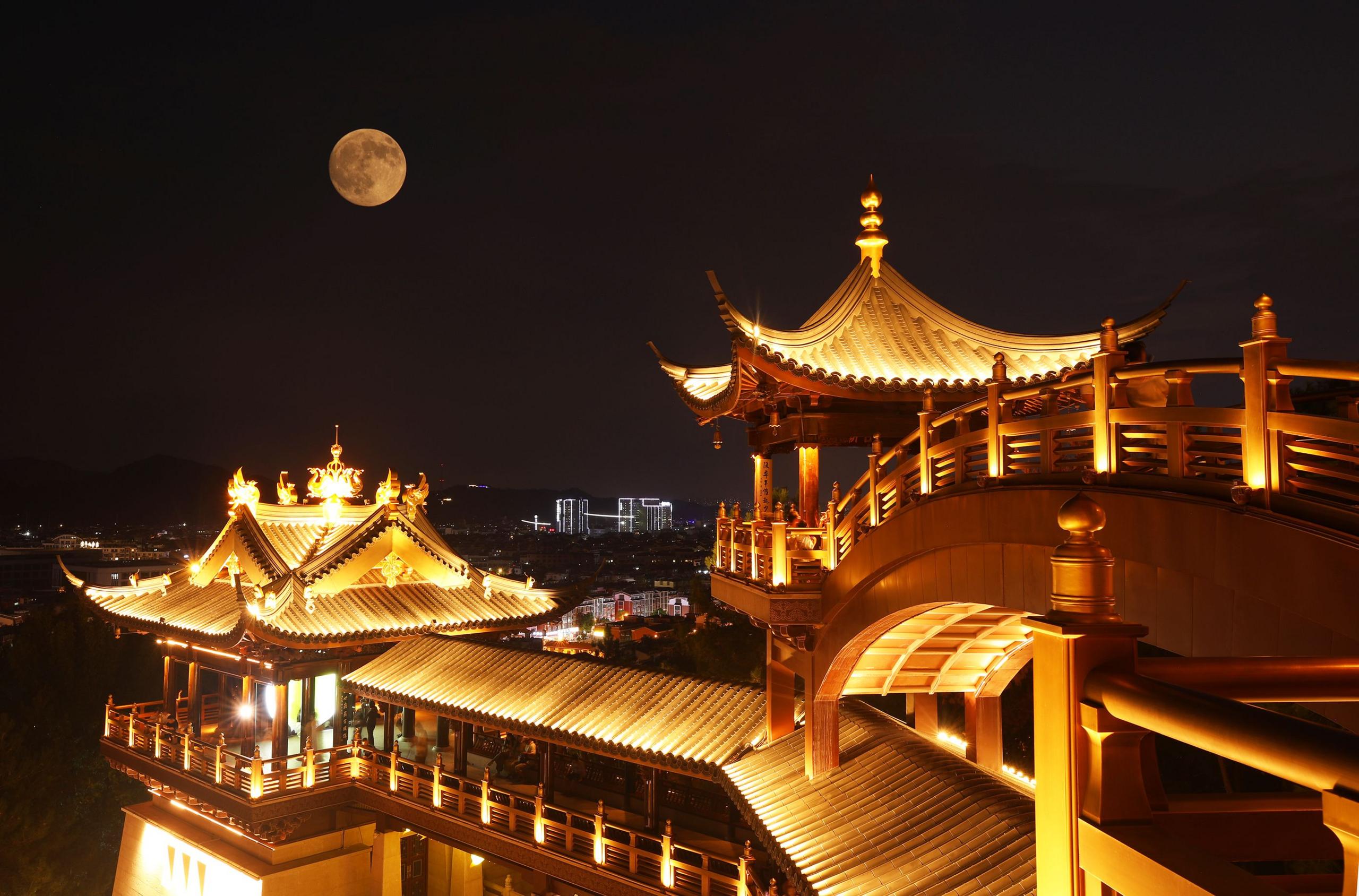 The full supermoon against a dark night sky, floating above a well-lit, ornately designed pavilion in Jinhua, Zhejiang Province.