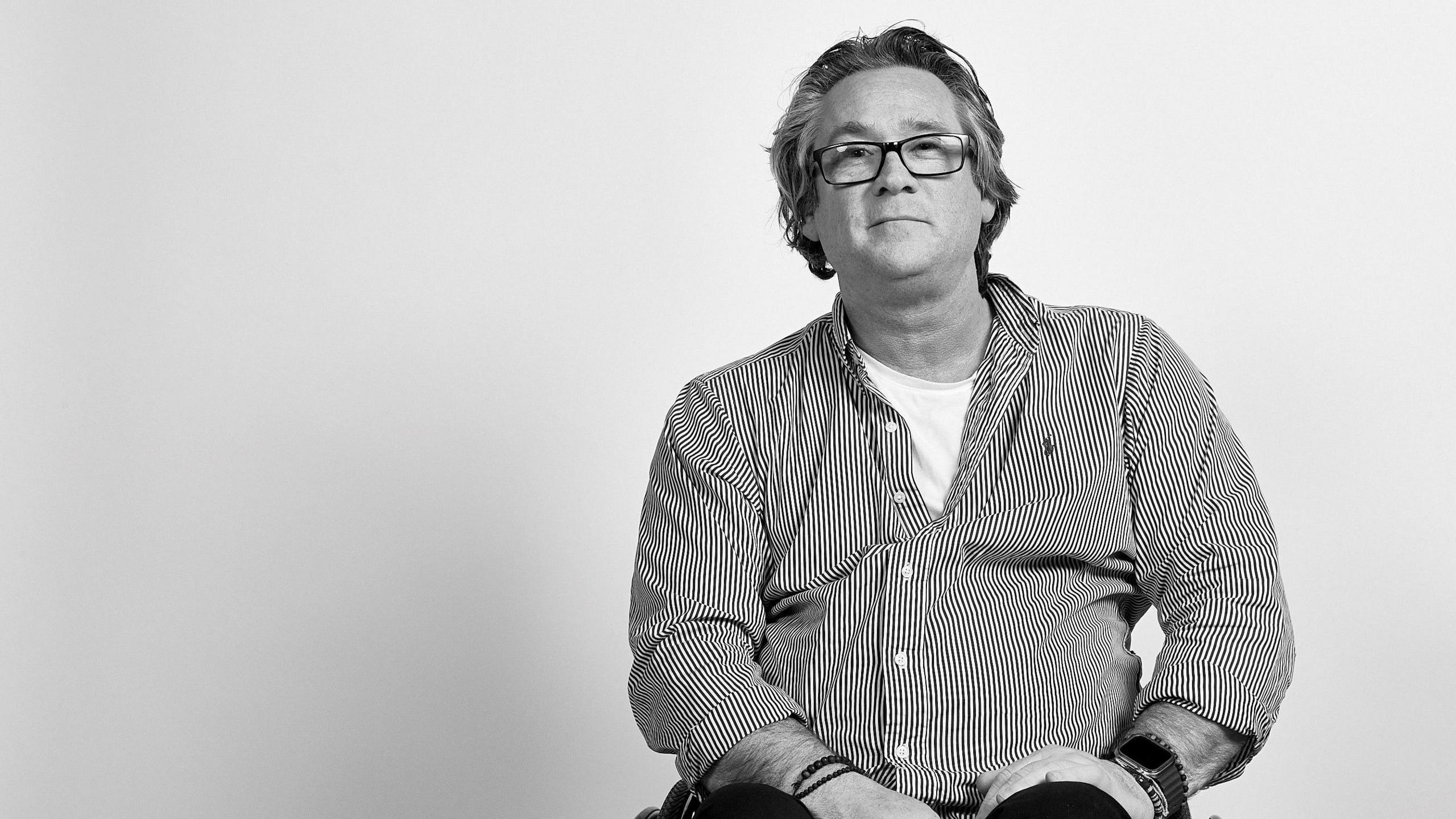 A black and white photo of a man with long, brushed back hair and glasses. he is sitting and wearing a stripy shirt with his hands resting on his lap