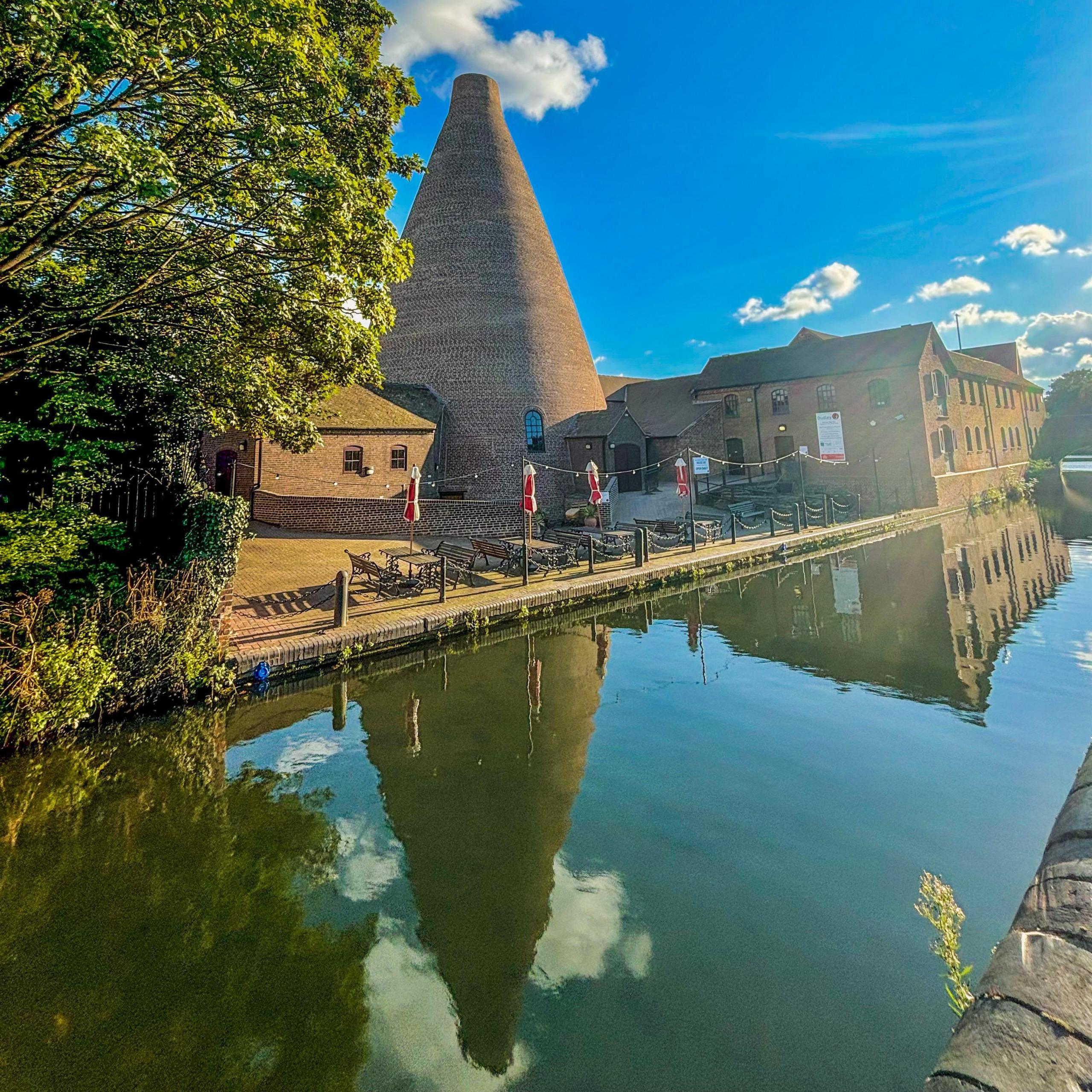 A red-brick, conical glass furnace is reflected in the adjacent canal. It is set among other historic industrial buildings with a tree encroaching on the shot from the left. 