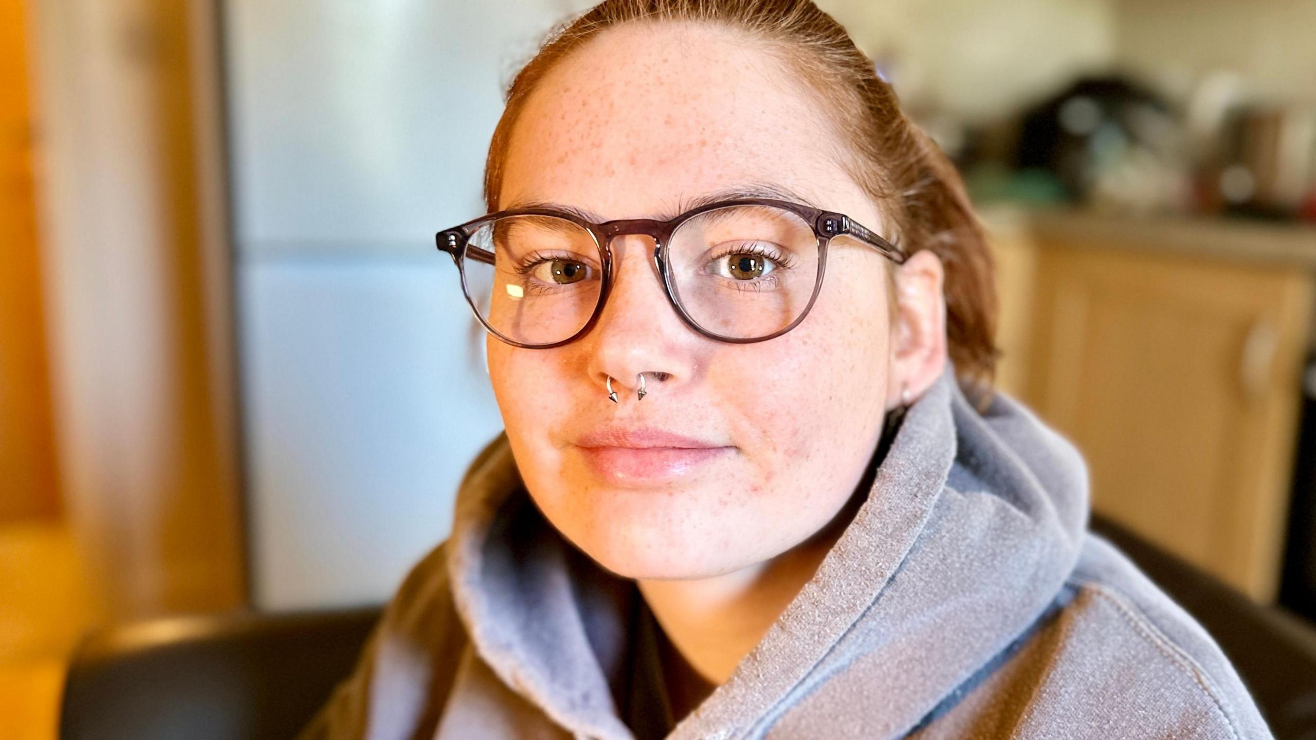 Head and shoulders portrait photograph of Stevie, taken in her kitchen, although the background is blurred. Stevie has auburn hair, tied back into a ponytail and brown glasses on. She has a nose piercing and is wearing a grey hoodie. 