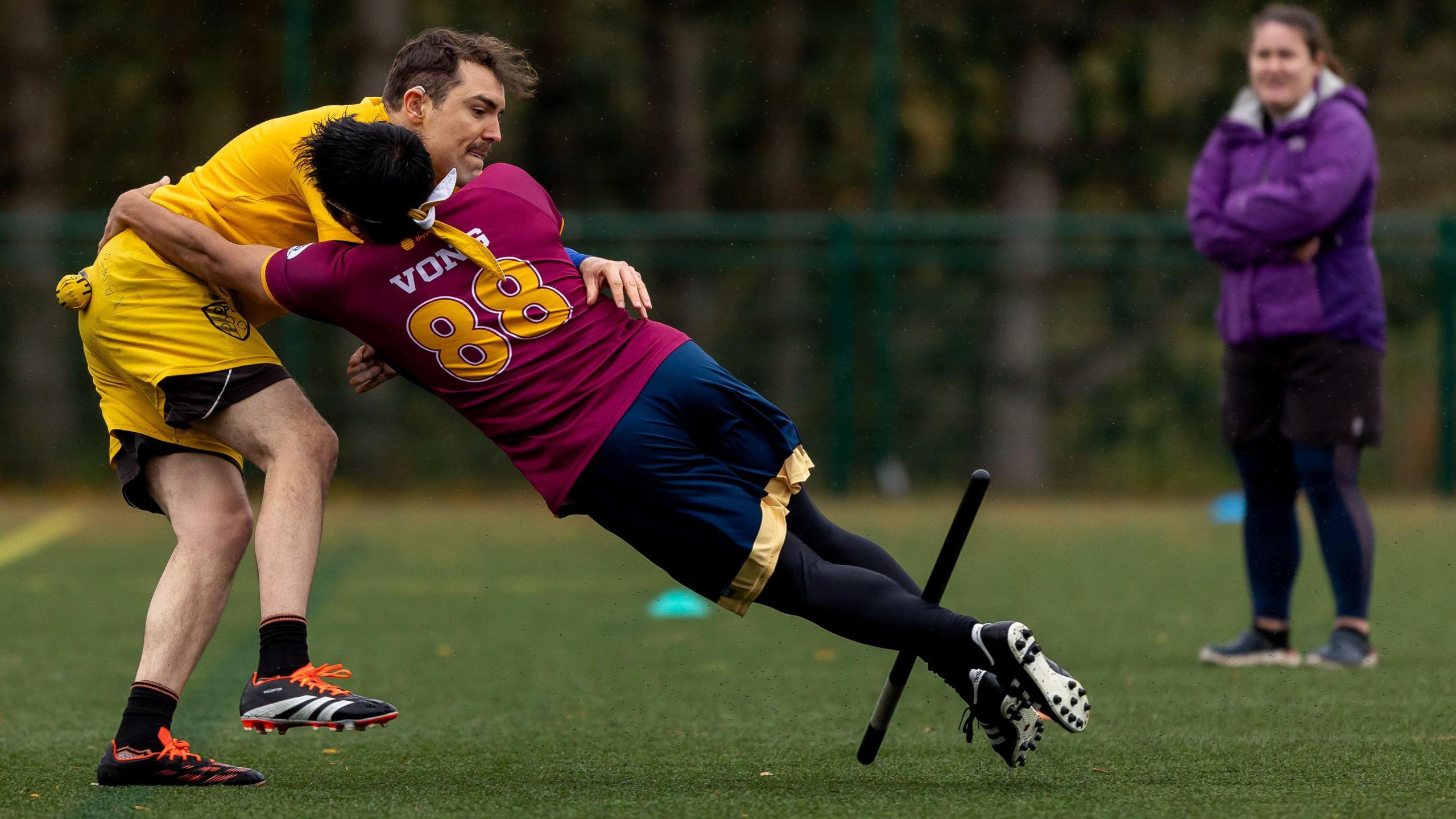 Two people in sports jerseys collide on a pitch. The player on the right grips a stick between their calves while attempting to grab around the other player with their arms. A yellow ball can be seen attached to the left player's shorts.