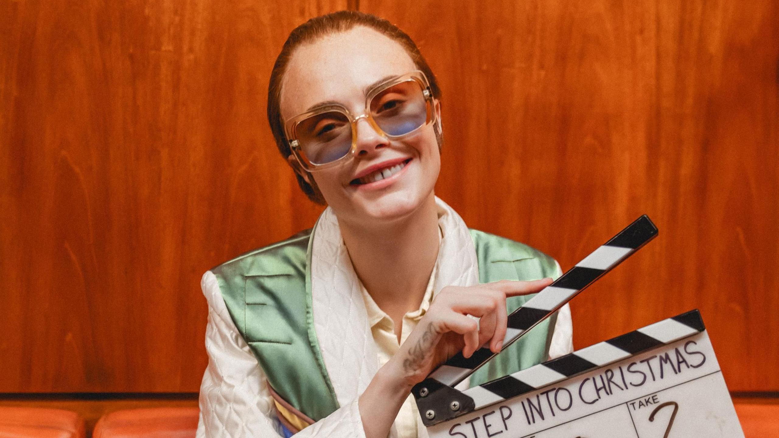 Cara Delevingne smiles the camera, dressed as Elton John. She wears a short, brown wig, large blue glasses, and a shiny green waistcoat. She is holding a clapperboard which says 'Step Into Christmas' on it.