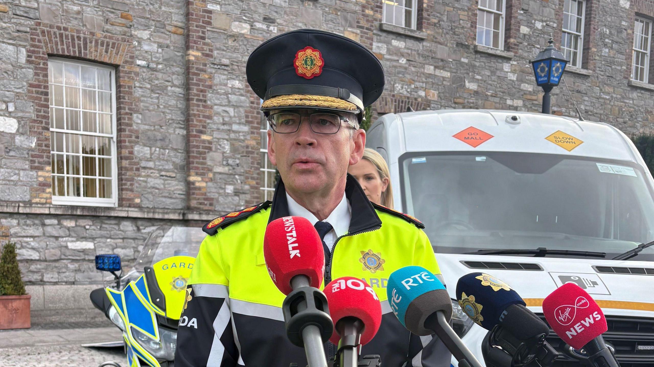 Garda Commissioner Drew Harris, wearing his Garda uniform, standing in front of a number of media microphones. There is a white van and a grey brick building behind him. 