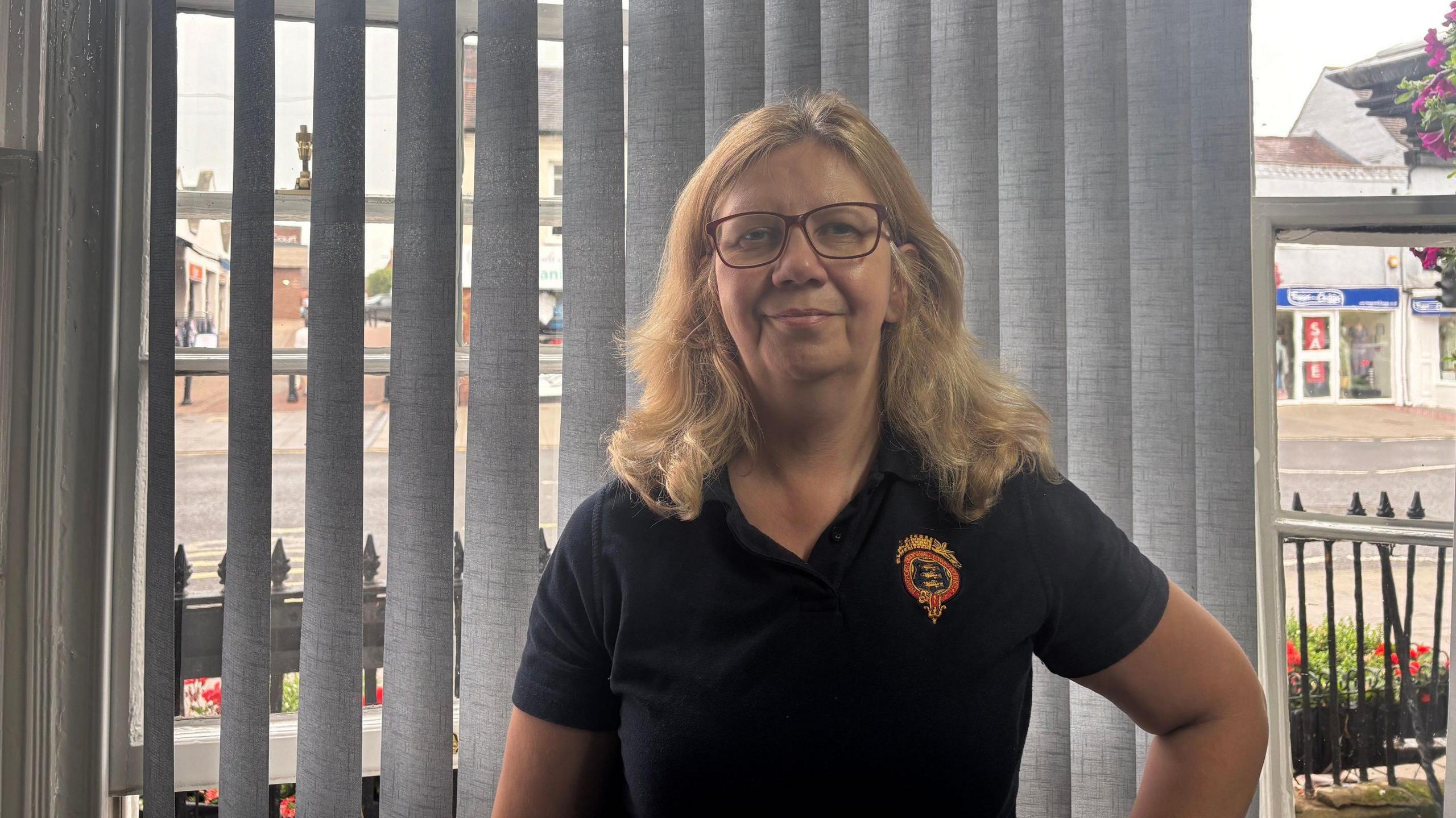 A woman with blonde hair and glasses is wearing a black polo top, standing in a building with blinds drawn behind her.
