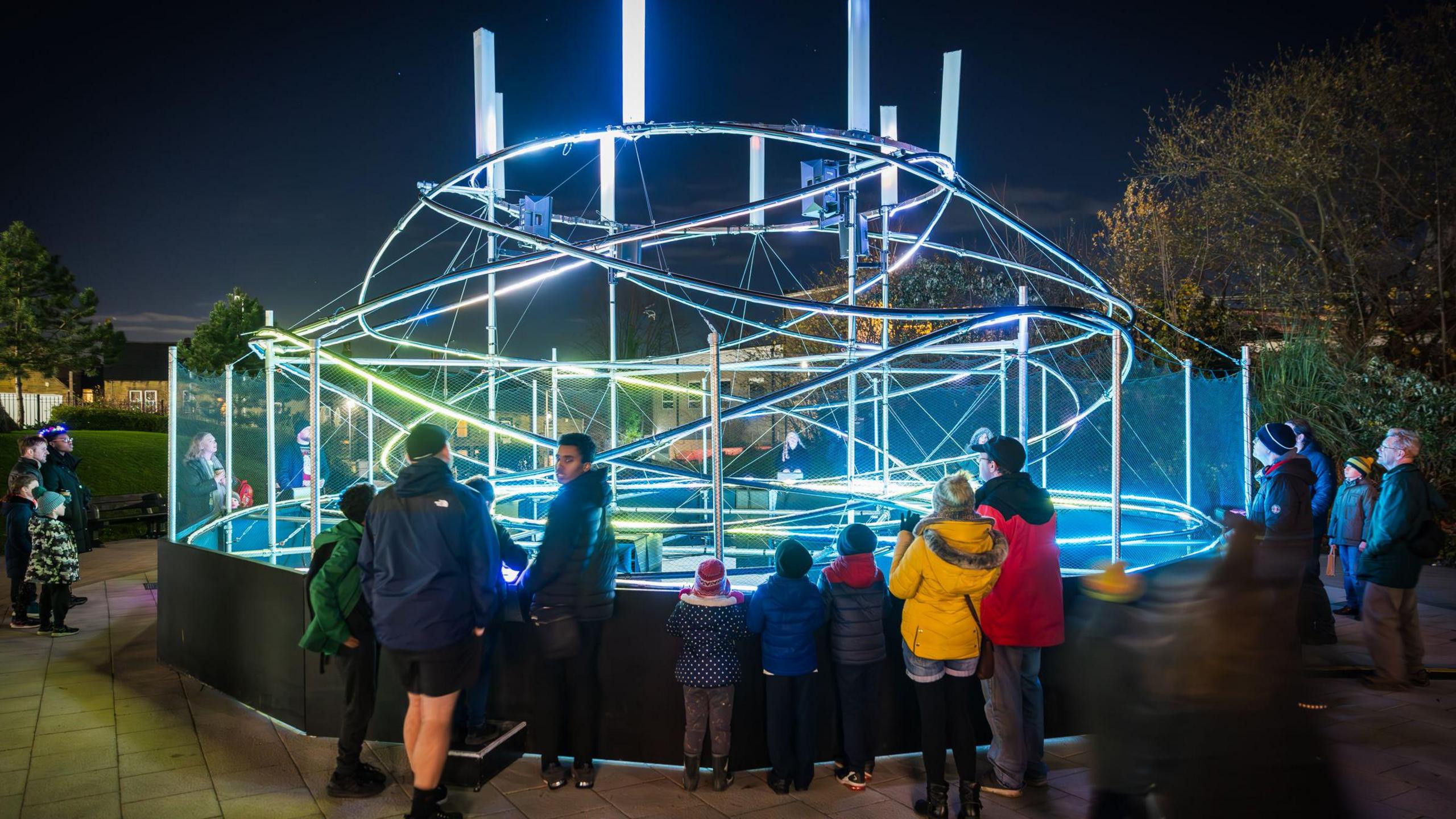 Around 20 people are standing around the edge of a large art installation made up of brightly lit tubes twisting around each other in a loose, haphazard spiral shape.