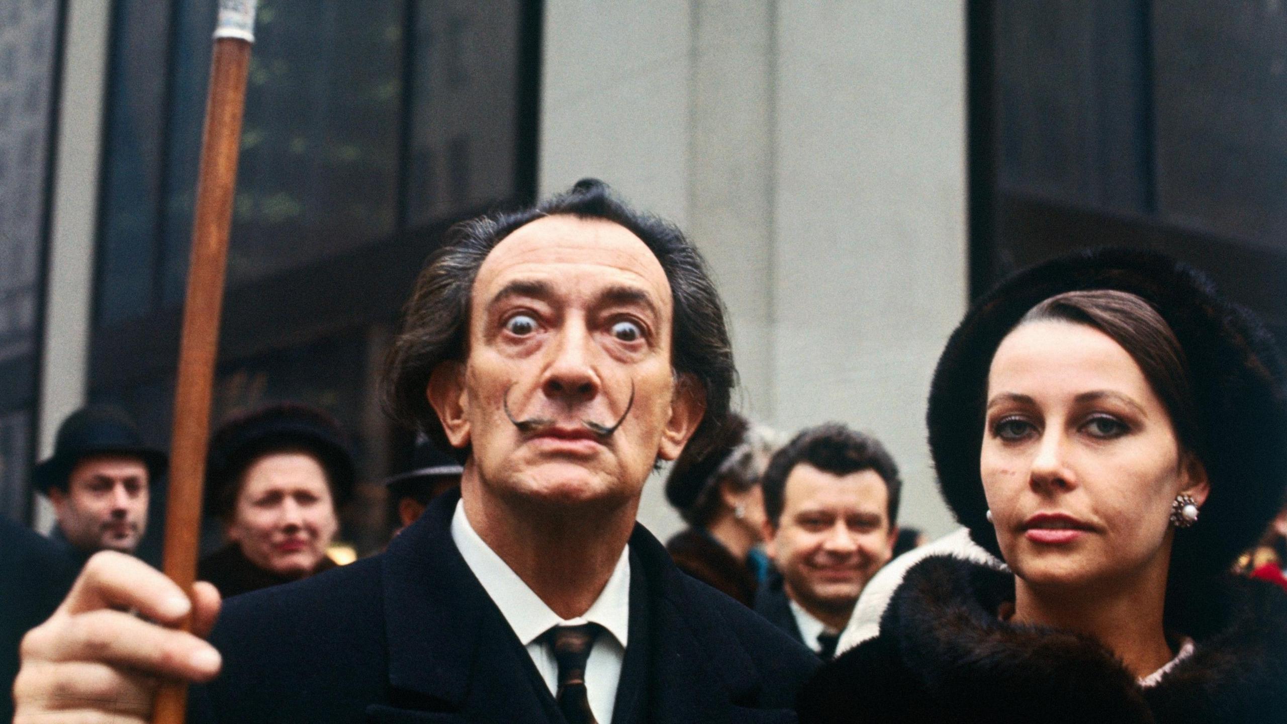 The artist Salvador Dali, wearing a dark suit and tie and white shirt, stares into the camera and lifts his walking cane with his right hand. He is standing next to a woman and in front of a large group of people, all wearing black.