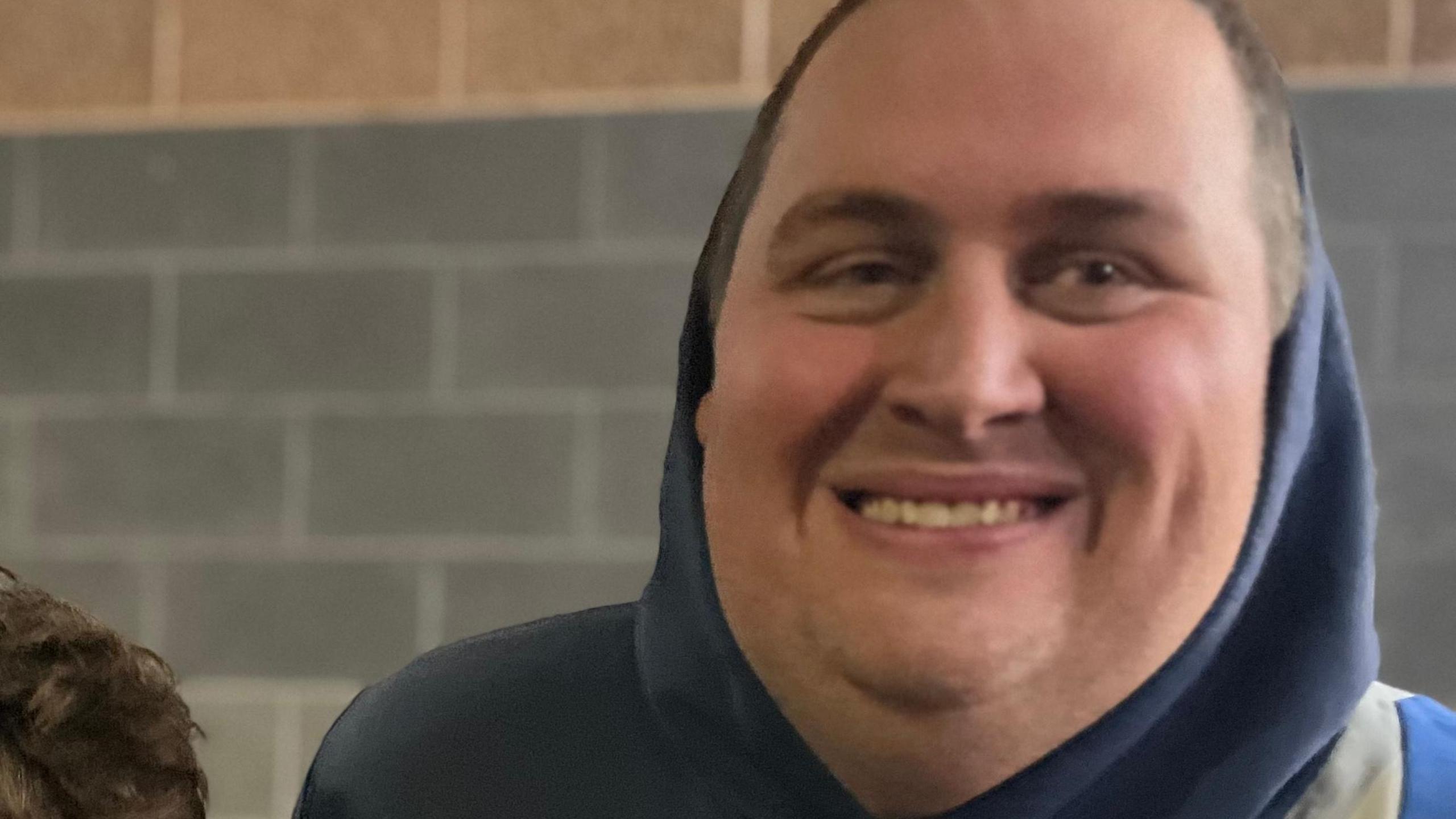 A close-up image of a man wearing a blue hoodie, the hoodie up. He is sanding in front of a grey brick wall