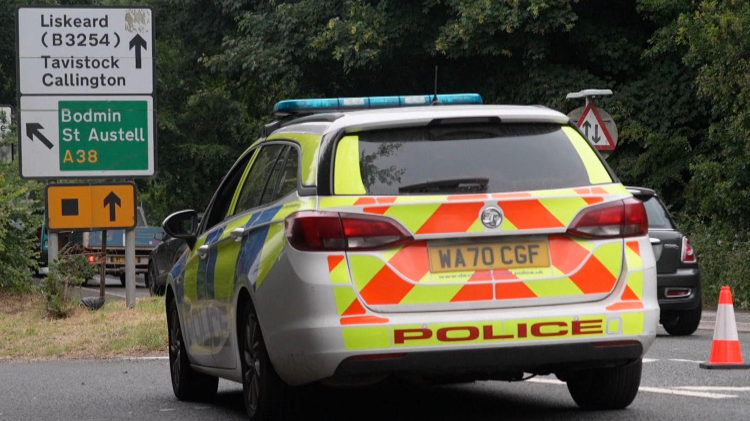 A police car is parked on a road. There is a road sign that reads: Liskeard (B3254) Tavistock Callington with an arrow indicating straight ahead. Under that it reads Bodmin St Austell A38 and points to the left. Under that sign is another sign that indicates a diversion route via a black box on orange background.