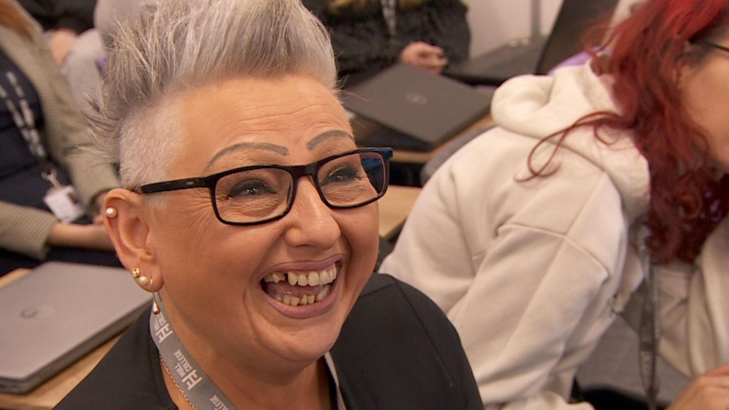 A woman with short grey, spikey hair and shaved eyebrows that have been pencilled in. She has dark rimmed glasses and has a huge smile for the camera. She is sitting in a classroom with other students using laptops.