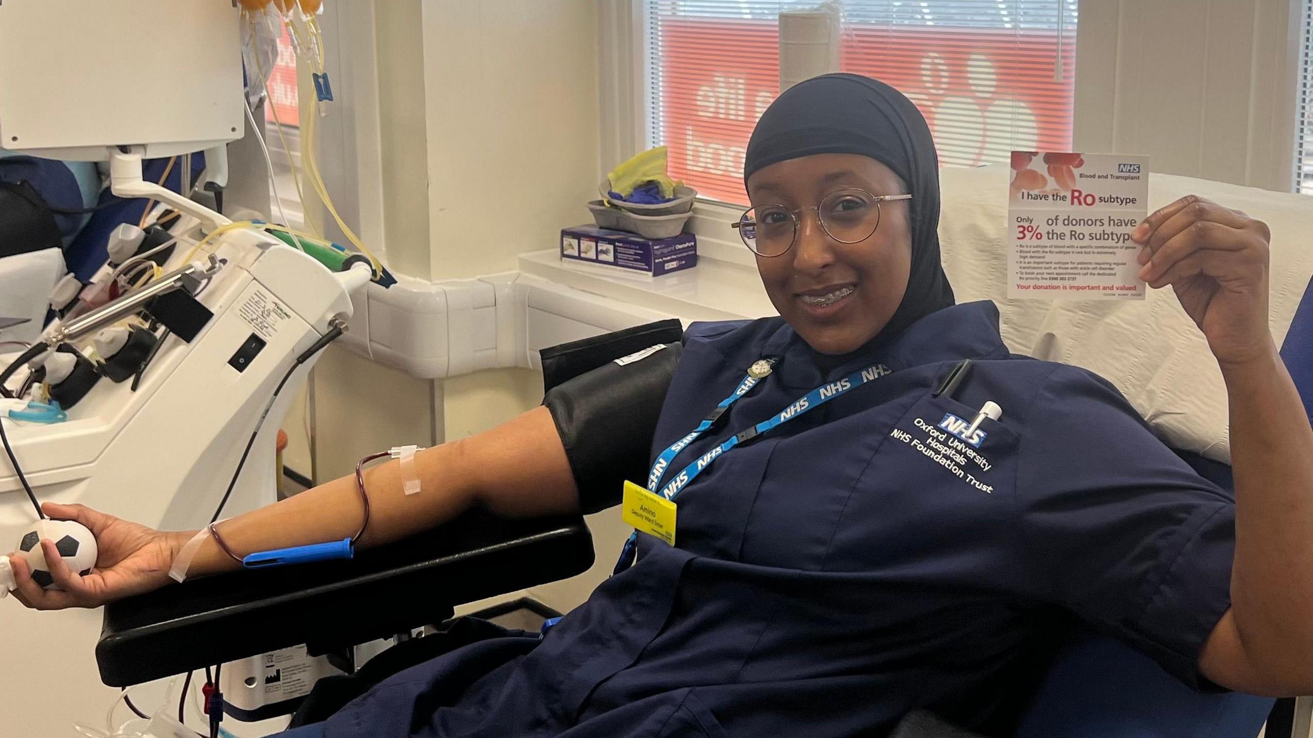 Ms Ali, still wearing glasses, a head covering and a dark blue nurse uniform,   smiles as she donates blood in the chair. She has a needle in her right arm, which also hold a football stress ball, and holds up a donation card to the camera with her other hand.