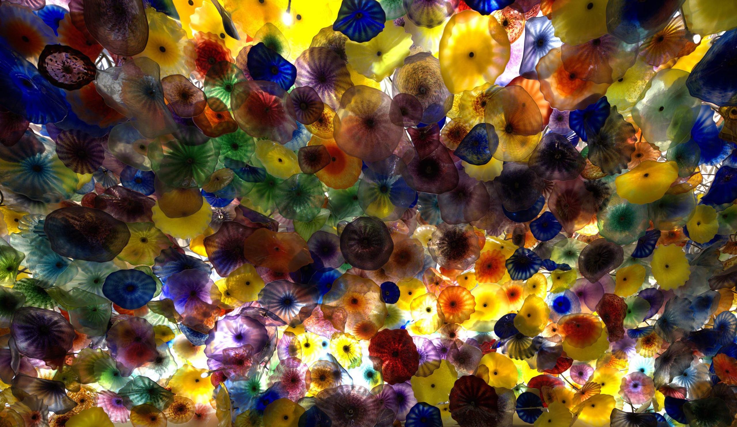 Multi-coloured glass flowers photographed from underneath. It looks like a sea of flowers in mostly yellow and purple with the sun peaking out from behind them.