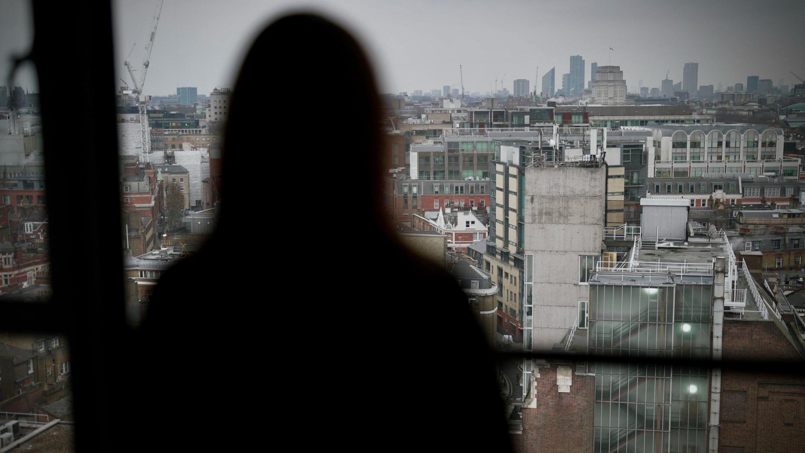 A silhouetted image of a woman, looking out from a high window over office blocks on an overcast day