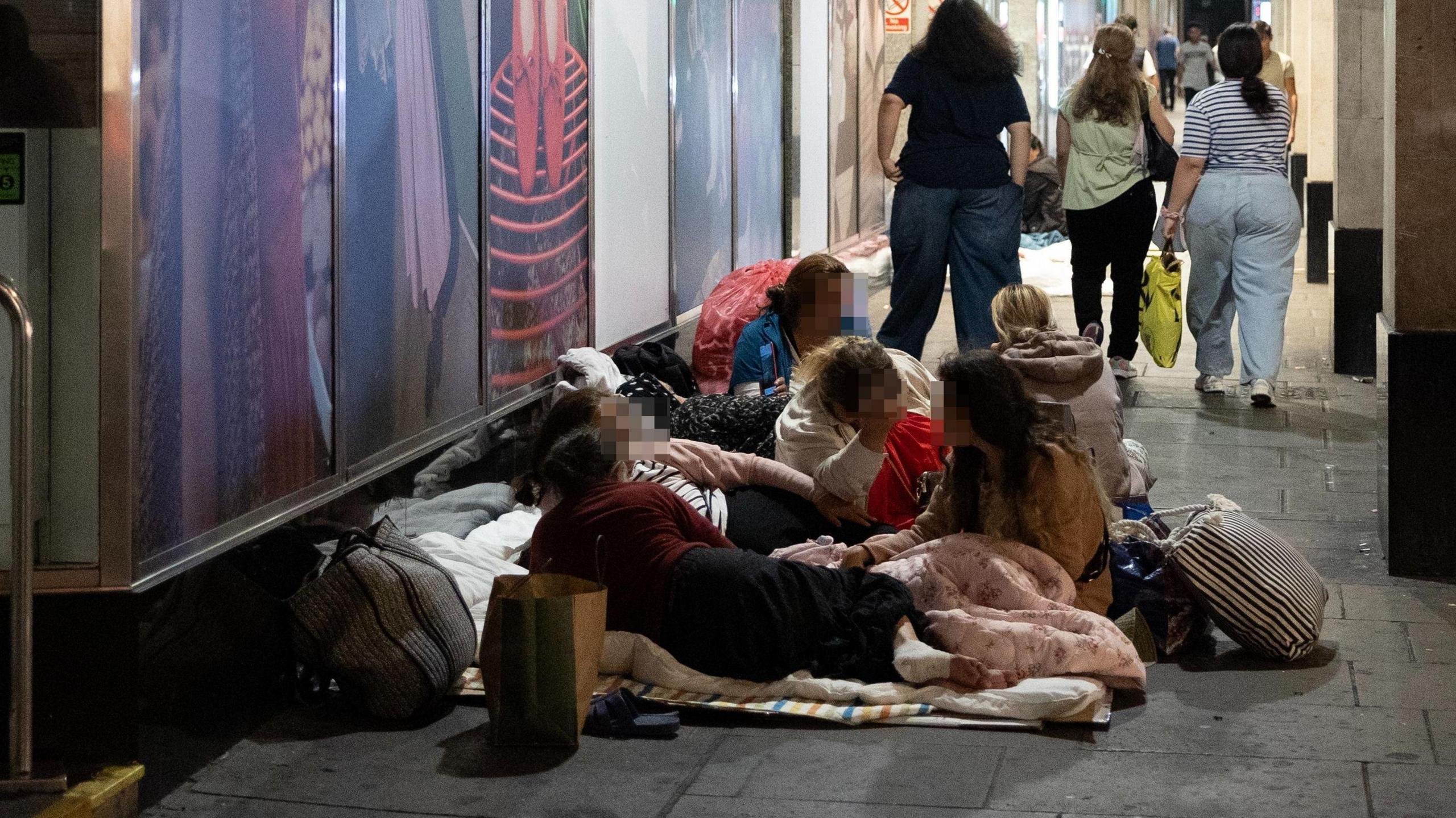 People lying down on blankets and pillows in front of a shop with fashion adverts in the building's windows. with large glass windows. A group walk past in the background.