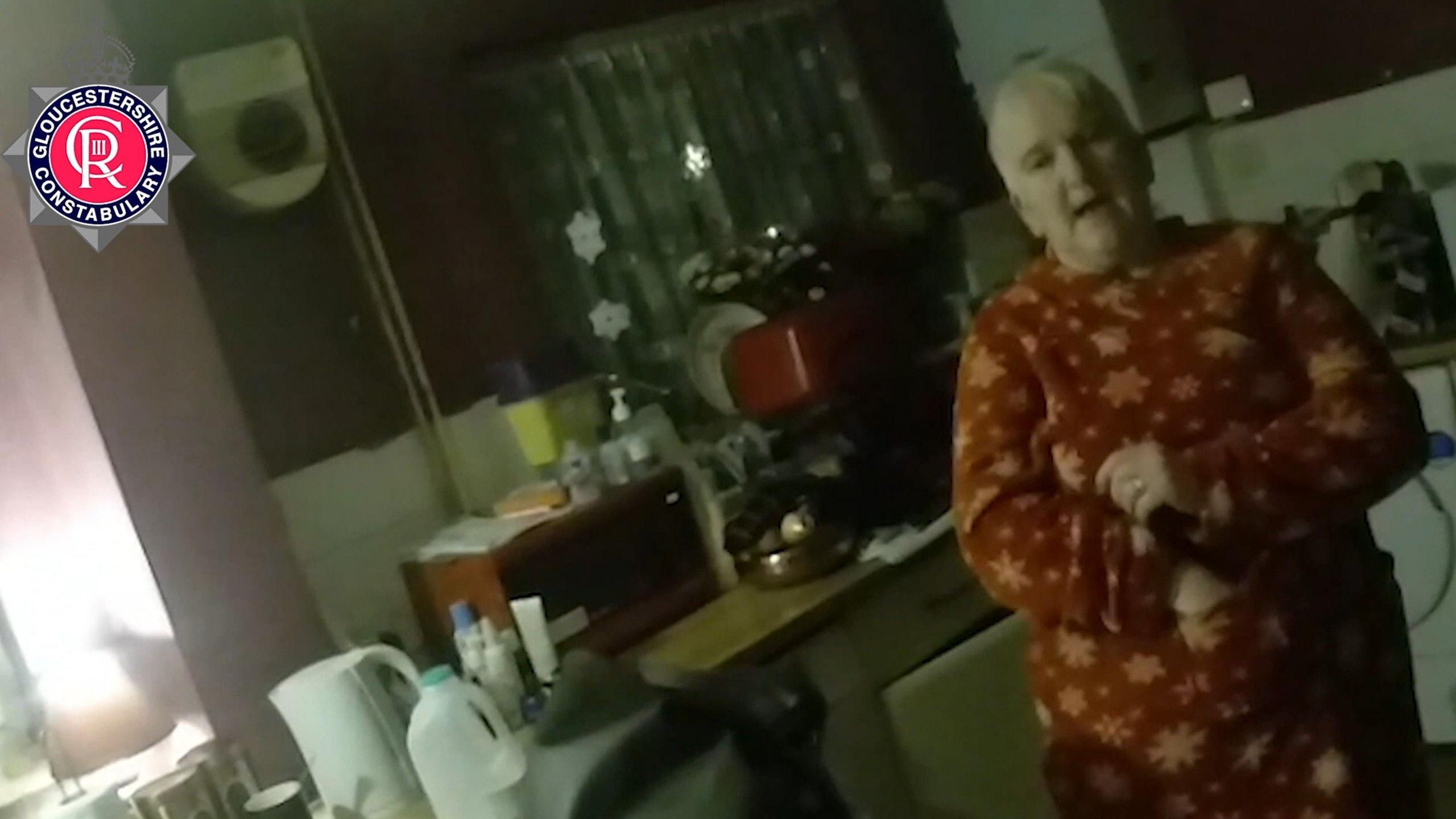Mandy Wixon stands in a kitchen in a red snowflake-pattern onesie. Behind her are various appliances including a kettle and microwave. 