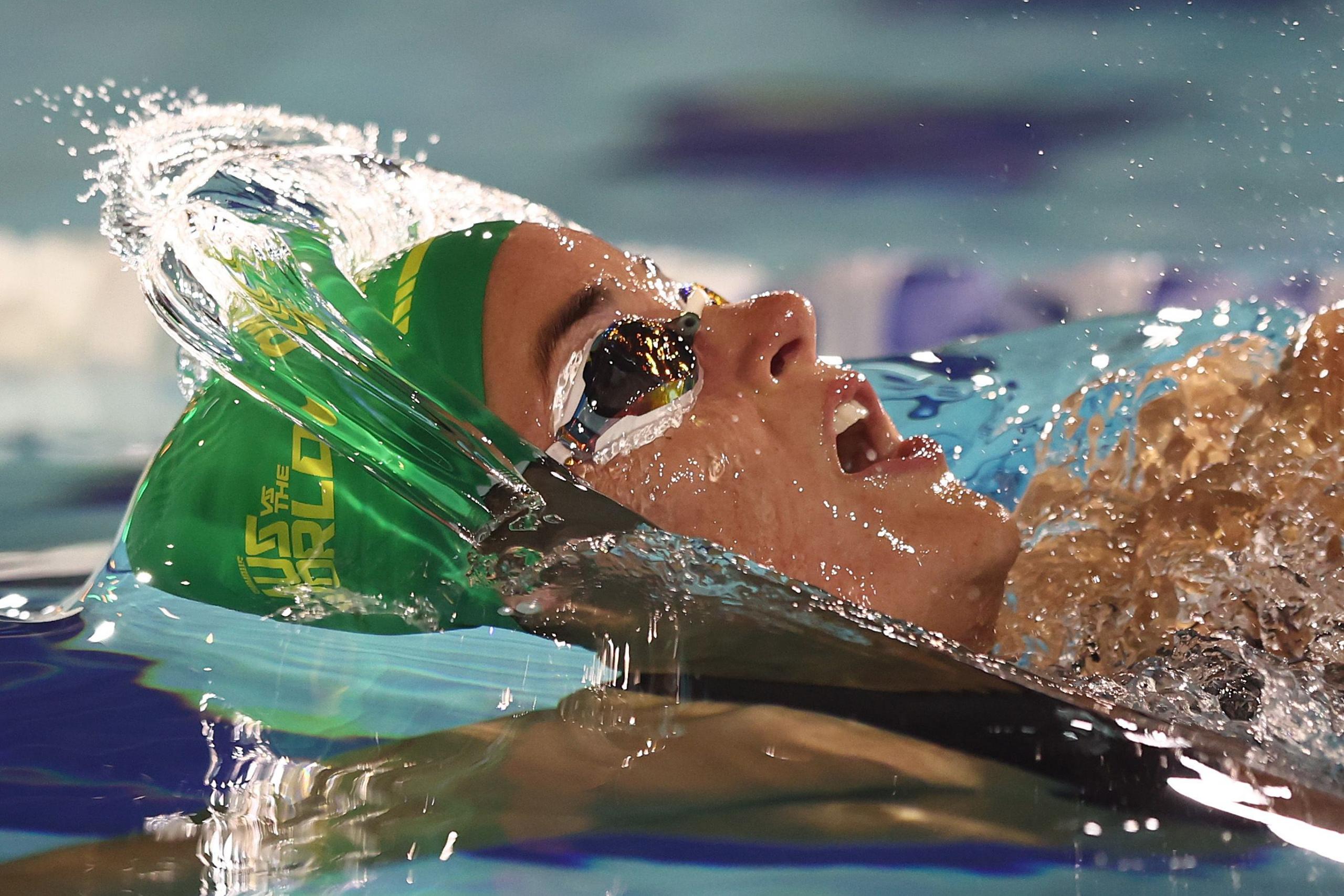 A swimmer wearing a green cap cuts through the water during a race, creating a splash that arcs upward as their arm drives forward along the surface of the pool.