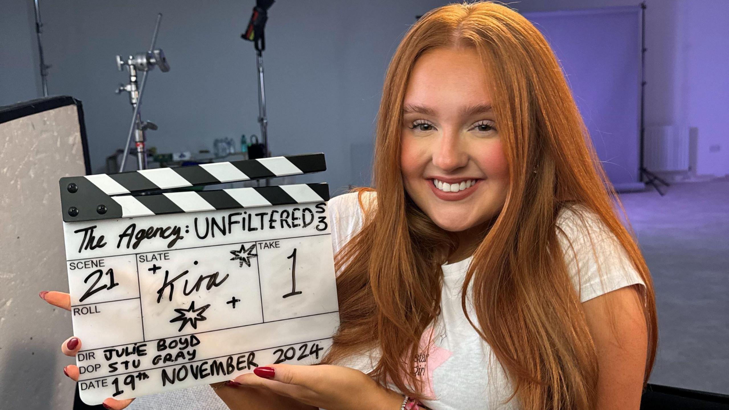 Kira McCaffery, wearing her long red hair down, and a white t-shirt, smiles broadly as she holds up a TV clapperboard.  She is sitting on a chair in a photographic studio with a backdrop and photography equipment behind her.