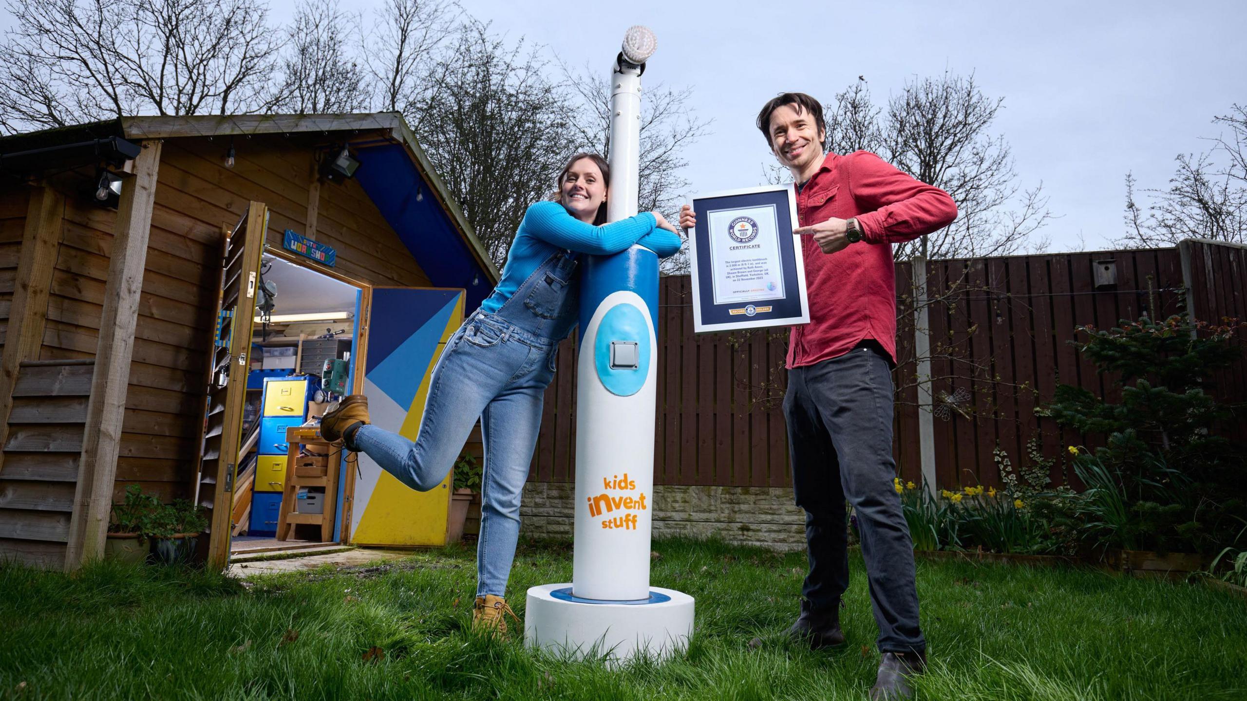 Ruth hugs the toothbrush, which is taller than her, while Shawn holds up the Guinness World Record certificate and points at it in delight. They are standing next to a shed in a garden