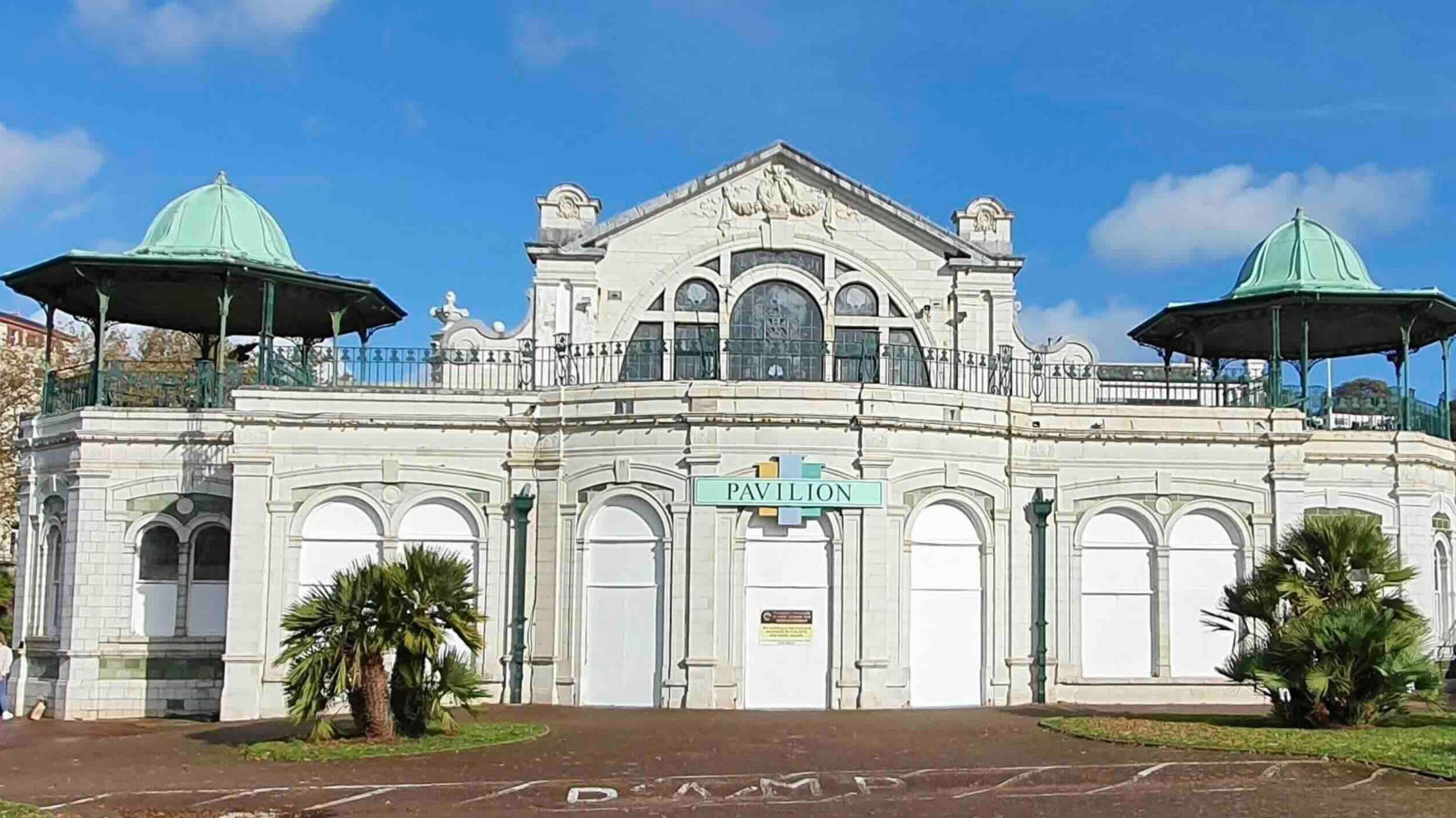 The Pavilion on Torquay seafront, a building faced with white tiles and topped with a pair of green domes. There is a sign over the door that says Pavilion