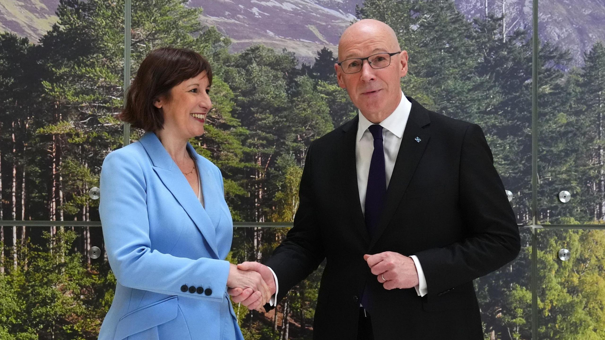 Rachel Reeves and John Swinney shaking hands in front of a wall covered by a picture of trees.