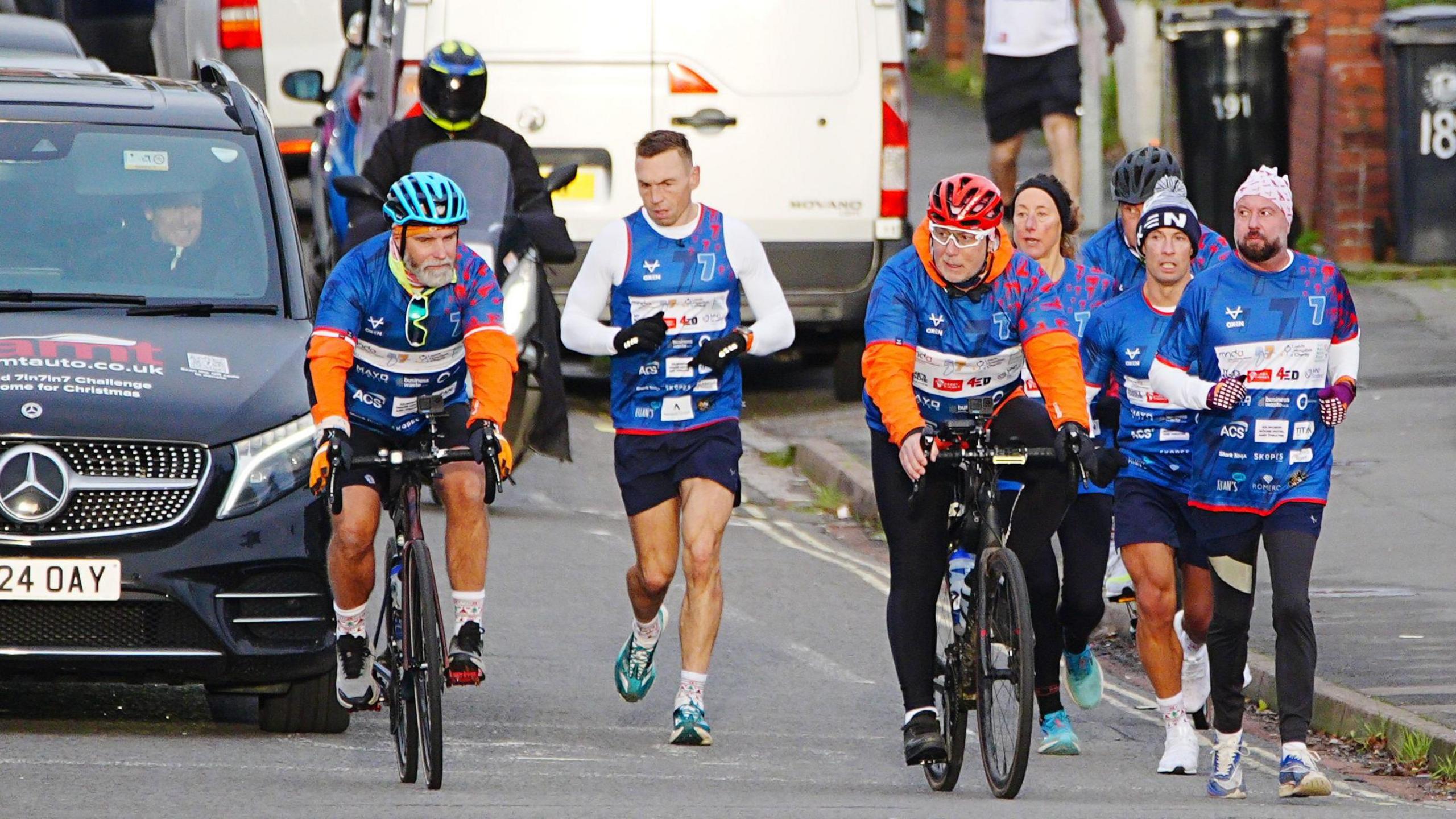 Kevin Sinfield and his support team running through residential streets. He is flanked by three people on bicycles and three other runners, while a support car trails behind.